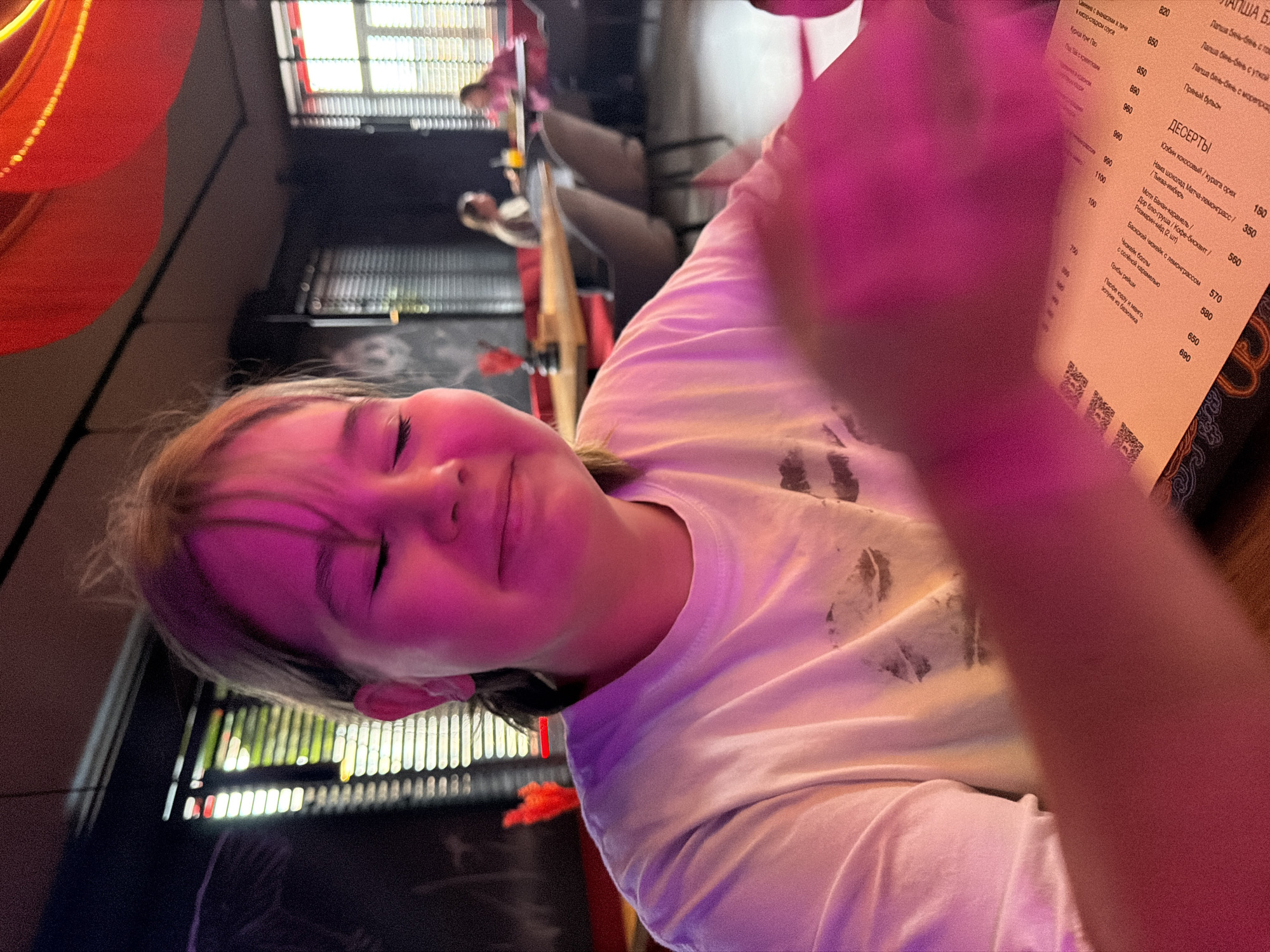 The media file captures a close-up, rotated portrait of a young person smiling with closed eyes, illuminated by strong pink and purple lighting. The main subject, likely a child or teenager, wears a white t-shirt and looks directly towards the camera. Their right hand is blurred in the foreground, also bathed in the pink light.
The scene is an indoor setting, likely a restaurant or cafe in Saint Petersburg, Russia, identifiable by the menu written in Russian. In the background, other out-of-focus patrons can be seen, along with architectural elements like dark grates, possibly part of windows or ceiling features. Red decorative elements, possibly lanterns, are visible in the top left corner.
The activity taking place appears to be dining, with the young person posing happily for a photograph. The lighting suggests an evening or low-light atmosphere.
Notable text visible on the menu includes "ДЕСЕРТЫ" (DESSERTS) followed by items such as "Юзбин кокосовый / курага орех" (Yuzbin coconut / dried apricot nut), "Моти Банан-карамель" (Mochi Banana-caramel), and "Баскский чизкейк с лемонграссом" (Basque cheesecake with lemongrass), along with corresponding prices in rubles (e.g., 180, 350, 560). Other dishes like "Свинина с ананасами и личи" (Pork with pineapple and lychee) and various "Лапша бянь-бянь" (Bian-bian noodles) options are also listed with prices. Several QR codes are also present at the bottom of the menu.
