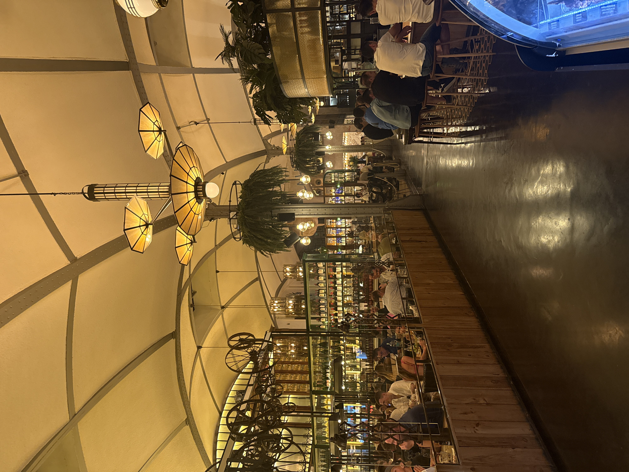 The image depicts the interior of a restaurant, likely in Barcelona, Spain, with a dimly lit ambiance. The ceiling is arched and covered with a textured material, supported by exposed beams and accented by industrial-style lighting fixtures. The restaurant appears to be a busy establishment, with numerous patrons seated at tables and at a bar area.
In the foreground, several individuals are seated at a bar, facing away from the camera. They are casually dressed, and their interactions are not visible. The bar itself is equipped with stools and what appears to be a display case on the right side of the frame.
The midground showcases more seating areas with patrons engaged in dining and conversation. The walls are lined with shelves displaying an extensive collection of bottles, suggesting a well-stocked bar or wine selection. Decorative elements like hanging plants and vintage industrial-style fixtures are visible, adding to the restaurant's unique aesthetic.
The background reveals more of the restaurant's interior, including additional seating and the continuation of the bar and shelving units. The overall impression is one of a lively and stylish dining and drinking establishment. The lighting creates a warm and inviting atmosphere, with reflections visible on the dark, polished floor.