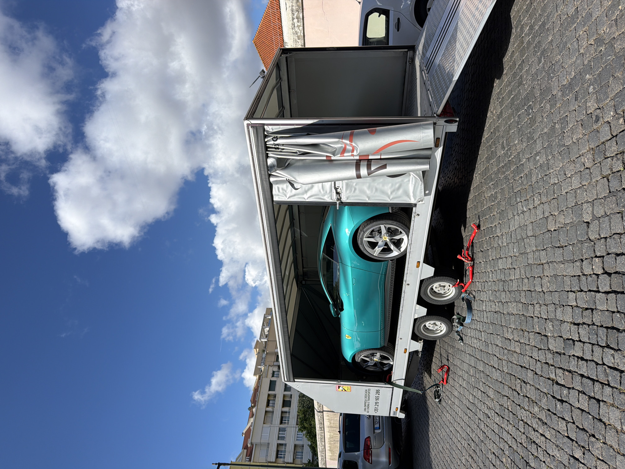 The image shows a blue Ferrari inside an open car carrier trailer parked on a cobblestone street. The location is Lisbon, Portugal, evidenced by the architecture in the background. The trailer has a silver-colored interior with a canvas covering. The sky is blue with white clouds. A silver-colored car is parked next to the trailer. Red straps and wheel chocks are visible around the trailer's wheels, securing the Ferrari.