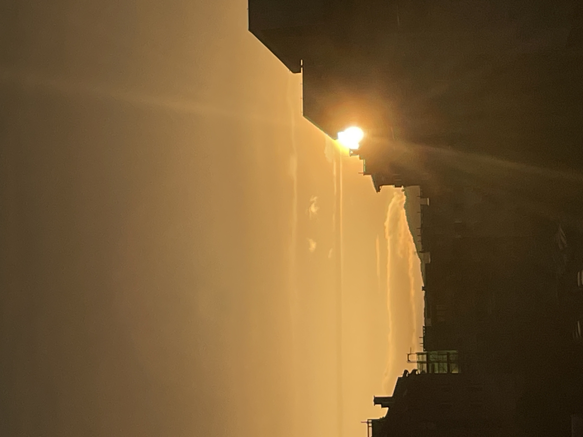 This image captures an urban scene in the city of Unknown, Unknown, at either sunrise or sunset. The sky, occupying the upper two-thirds of the frame, displays a gradient of warm golden-orange hues, with a few small, diffuse clouds visible across the horizon and in the mid-left. The sun, a brilliant white-yellow light source, is partially obscured by the dark silhouette of a multi-story building on the right, generating a prominent light flare and upward-stretching crepuscular rays. The lower portion of the image features a contiguous silhouette of buildings, appearing dark brown to black against the bright sky. On the far right, a building exhibits a stepped roofline. To its left, a row of residential or commercial structures includes visible chimneys and a distinctive, partially illuminated, scaffolding-like structure in the mid-ground. In the distant background, between the building silhouettes and the clouds, the outline of hills or mountains is faintly discernible. No people are visible.