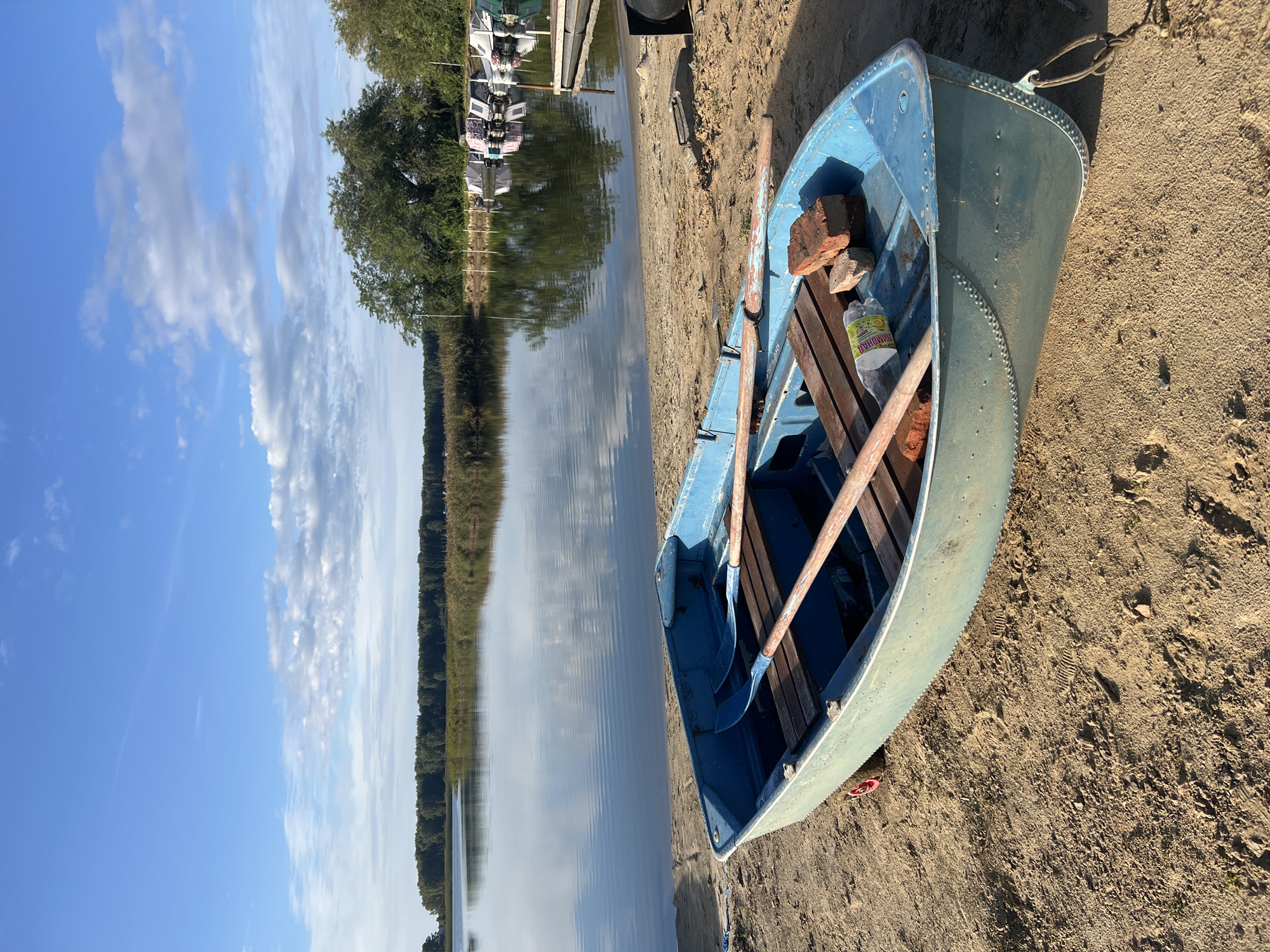 The image depicts a blue rowboat resting on a sandy shore next to a calm lake. The boat contains two oars, a plastic bottle, and two bricks. In the background, several other boats are docked on the water, and a dense line of trees forms the horizon. The sky is blue with scattered white clouds. The location is the shore of the lake in Novopichugovo, Russia.