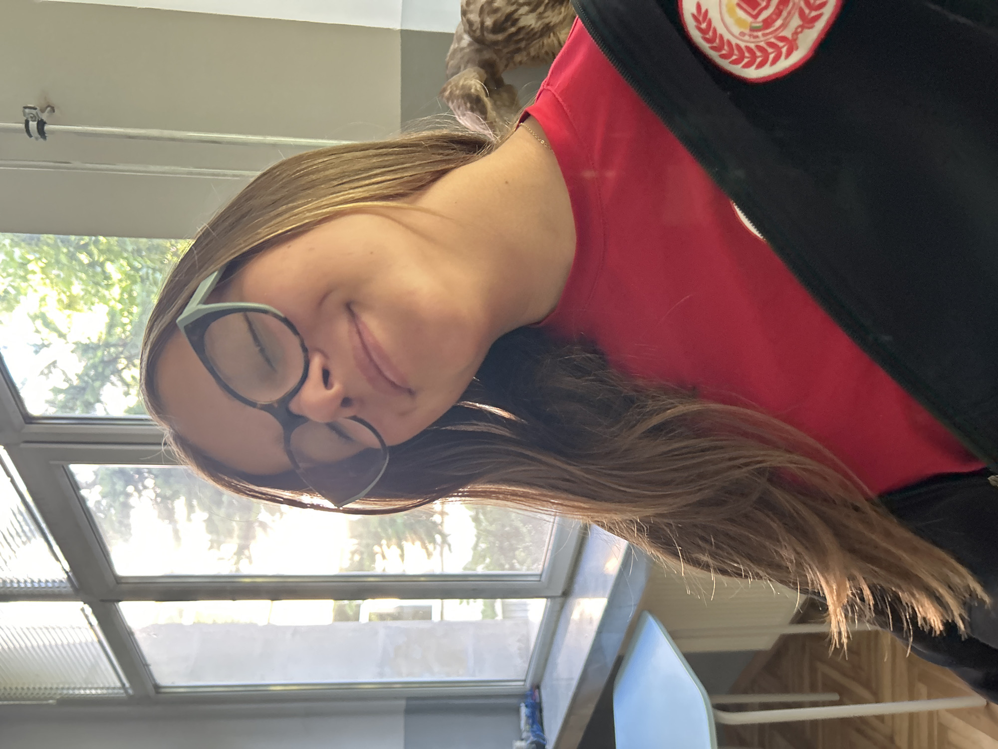 The image, oriented correctly, features a young woman smiling contentedly with a closed-mouth expression. She has long, straight light brown hair and wears black-framed glasses with a distinctive light blue-green accent on the upper part of the frame. Her attire includes a bright red top layered under a dark, possibly black, jacket. A circular patch, predominantly red and white with a red crest design, is visible on the right shoulder area of her jacket, suggesting affiliation with an institution or organization.
The woman is indoors, with a large, multi-pane window directly behind her. Bright daylight streams through the window, illuminating green trees and foliage outside, indicating a sunny daytime setting. Inside, parts of a light-colored table or desk and a light blue chair are visible on the right side of the frame. Above the woman, a white ceiling or upper wall features a metal track or pipe with a small clamp. A blurry, textured brown object is partially visible behind her head.
The setting appears to be an educational environment, such as a classroom or common area, potentially in Sofia, Bulgaria, as indicated by the context. The woman is posing for a photograph, conveying a cheerful and pleasant mood. No visible text is present in the image, only the graphical emblem on her jacket.