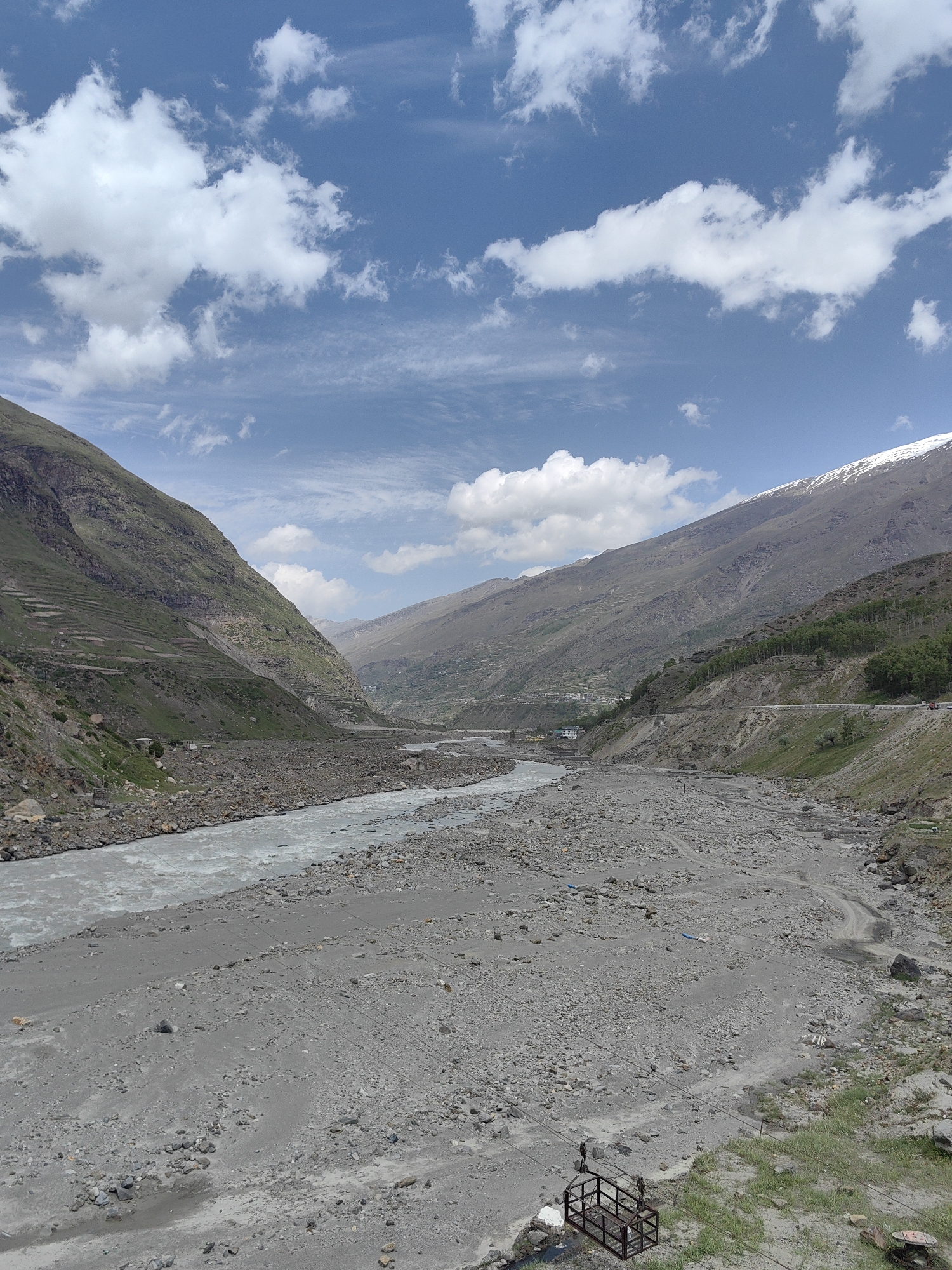 The image depicts a wide, rocky riverbed in the vicinity of Sissu, India. A turbulent, grey river flows through the left side of the frame, bordered by steep, green-covered mountains that display terraced farming on their slopes. The riverbed itself is composed of a vast expanse of grey gravel and stones, with a few scattered patches of green vegetation. In the distance, the mountains continue, with one peak showing a dusting of snow. The sky is a clear blue, dotted with fluffy white clouds. A road snakes along the side of one of the mountains in the middle ground, with a few vehicles visible. In the foreground, a rusted metal cage, likely part of a cable car system, rests on the rocky ground.