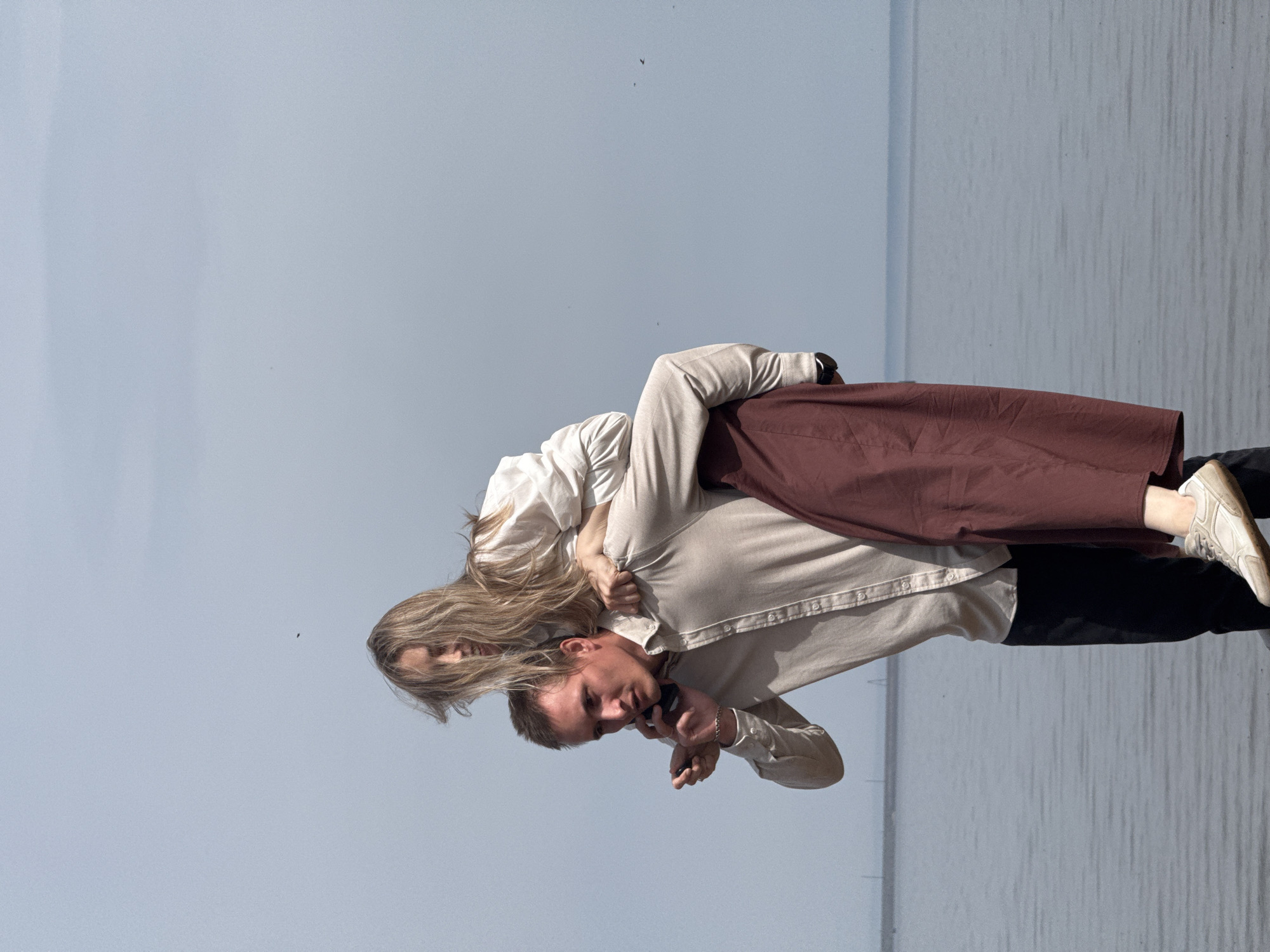 A man is giving a woman a piggyback ride on a body of water in the city of Chaikovsky, Russia. The man wears a tan, button-down shirt, black pants, and white sneakers. The woman wears a white, button-down shirt, and a long, brown skirt. The man is talking on a cell phone. The background shows a grey sky and water.