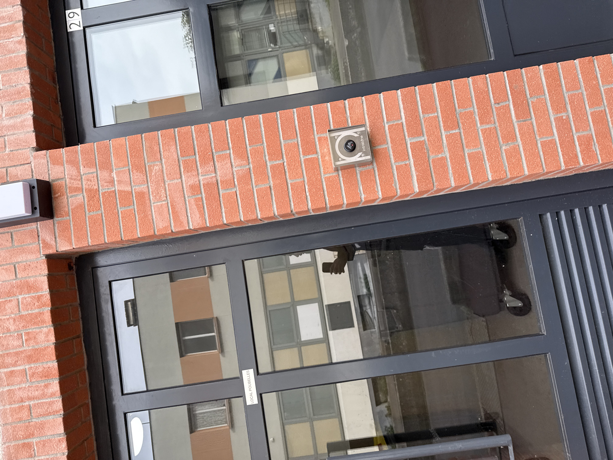 An eye-level, medium-shot photograph shows the exterior of a brick building in Domont, France. The building is constructed of reddish-brown bricks arranged in a running bond pattern. A dark gray, rectangular metal frame surrounds two glass windows. The top window displays the number "29" in the upper left corner. Reflected in the lower window is another building with gray and beige walls. A sticker is visible on the lower window, and a metal callbox is mounted on the brick wall between the two windows. The reflection in the lower window also shows a person holding a phone. A rectangular white light fixture is on the left edge of the image.