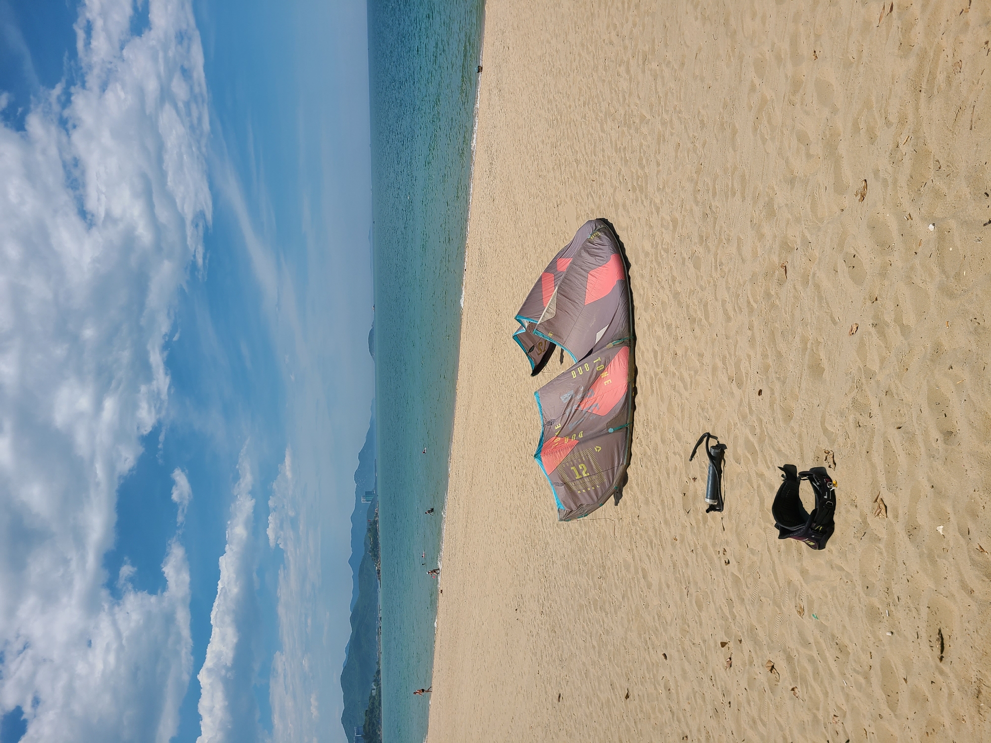 This media file depicts a tropical beach scene in Nha Trang, Vietnam. The image is oriented vertically, with the sky and ocean occupying the left portion and the sandy beach the right. The sky is a vibrant blue, dotted with scattered white clouds. The ocean is a clear turquoise, with gentle waves lapping the shore.
On the sandy beach, the primary subjects are two pieces of kiteboarding equipment: a kite and a harness. The kite is large and colorful, with shades of grey, pink, and black. It appears to be folded or partially deflated and lies on the sand. Next to it is a black kiteboarding harness, also resting on the beach. A control bar, likely for the kite, is also visible.
In the distance, along the shoreline, several small figures of people can be seen enjoying the beach and the water, some wading or swimming. The backdrop of the scene includes lush green mountains that rise behind the coastline, characteristic of the Nha Trang region. Buildings are faintly visible nestled against the foothills.
The lighting suggests it is daytime, likely mid-morning or afternoon, with bright sunlight casting subtle shadows on the sand. The weather appears to be clear and pleasant, ideal for beach activities. The overall mood is one of leisure and outdoor recreation. Visible text on the kite includes the number "12" and what appears to be a brand logo.