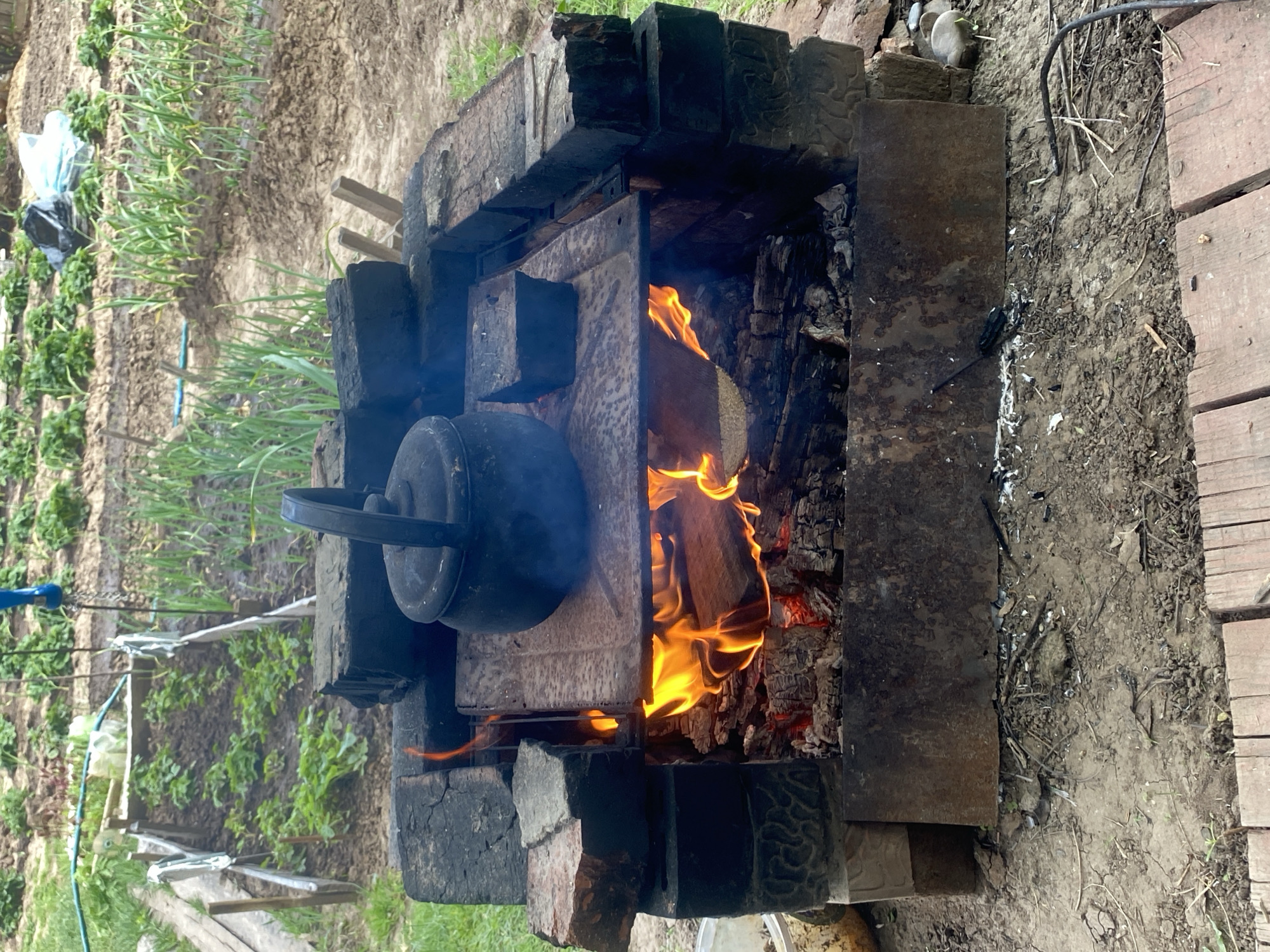 A kettle sits atop a brick-and-metal fire pit in an outdoor setting. Flames are visible within the pit. To the left, a garden is present. The image was taken in the city of Прикамский, Russia. A wooden structure is at the edge of the picture.