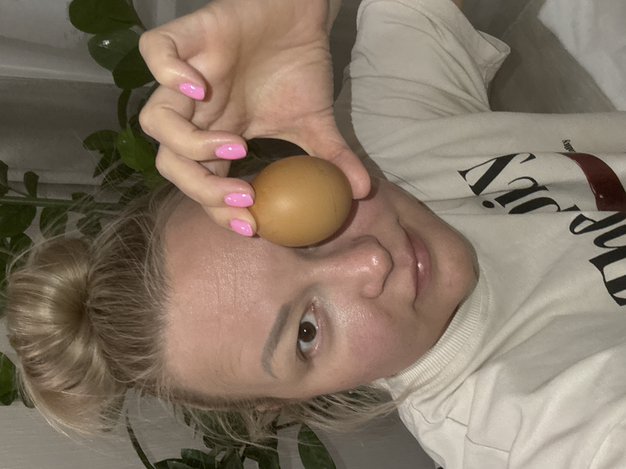 A woman lies on a light-colored surface, possibly a bed or couch, with a plant visible on the left side of the frame. Her blonde hair is pulled back in a bun. She holds a brown egg in her hand near her forehead, fingers visible with pink nail polish. She is wearing a light-colored top with black and red lettering. The image does not contain enough information to confirm it was taken in Izhevsk, Russia.