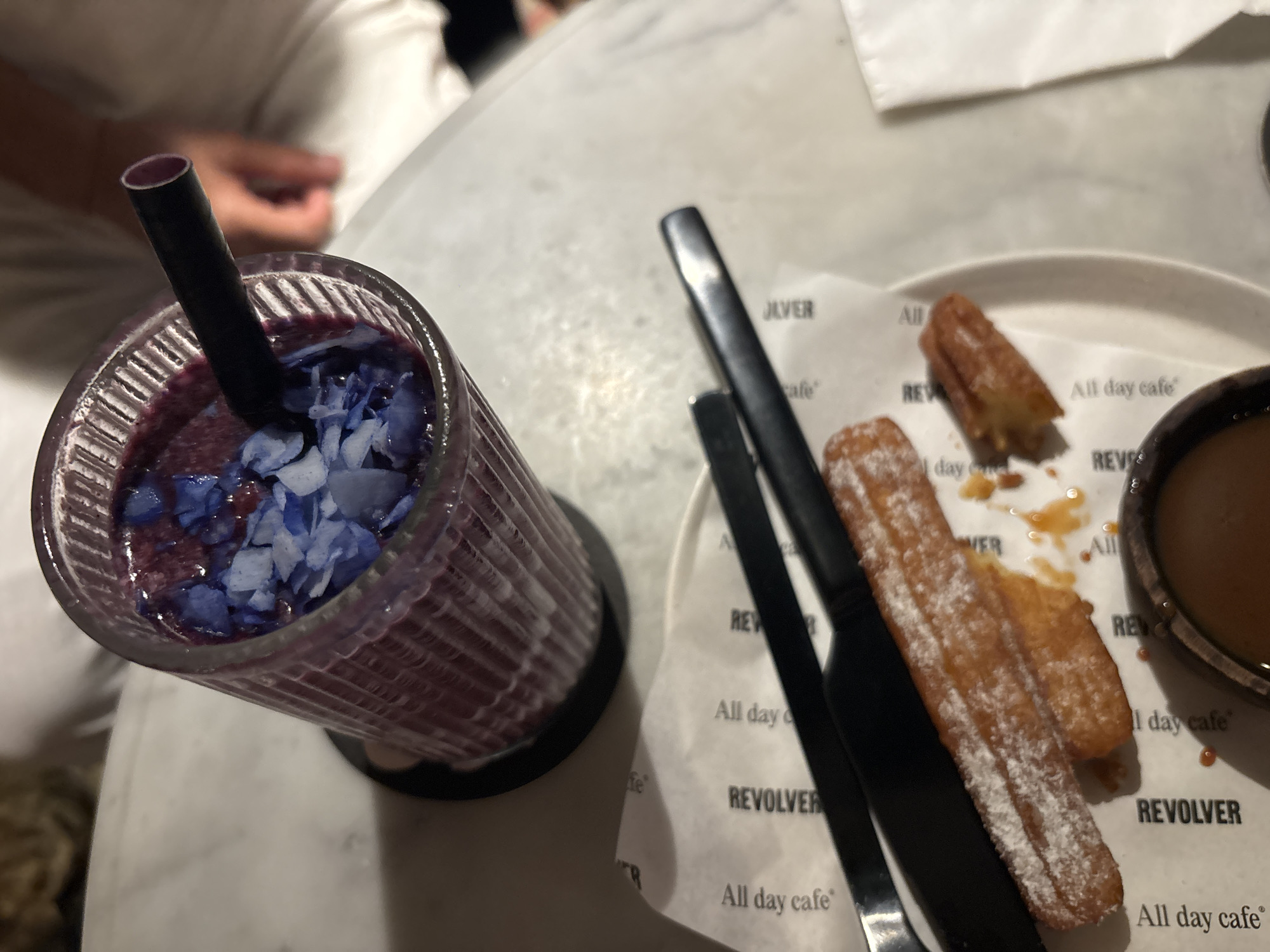 A close-up, high-angle shot shows a glass of purple smoothie with blue petals and shredded coconut, alongside churros dusted with powdered sugar and a ramekin of caramel sauce on a table at Revolver Espresso, North Kuta, Indonesia.