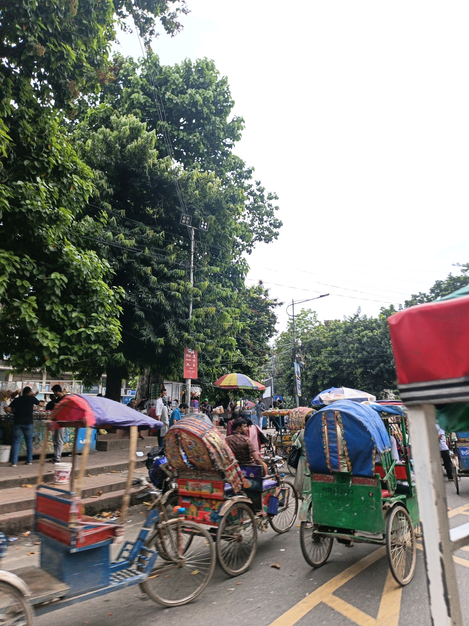 The image depicts a busy street scene in Dhaka, Bangladesh, characterized by a dense urban environment with lush trees lining the road. The foreground is dominated by several cycle rickshaws, the iconic mode of transport in the city. One rickshaw, painted blue and red with a purple canopy, is prominently featured, showing signs of use with a weathered appearance. Its seating area is clearly visible. To the right, another rickshaw, predominantly green with a blue canopy, is also visible, with intricate patterns adorning its fabric. Several people are present, some riding the rickshaws and others walking or standing by the roadside. One person, dressed in a brown patterned shirt, is seated in a rickshaw. A yellow and red umbrella is open, providing shade. The background reveals more of the city's infrastructure, including buildings and electrical wires suspended from poles, indicating a typical urban setting. The road surface displays yellow lane markings. The overall impression is one of vibrant, everyday street life in Dhaka.