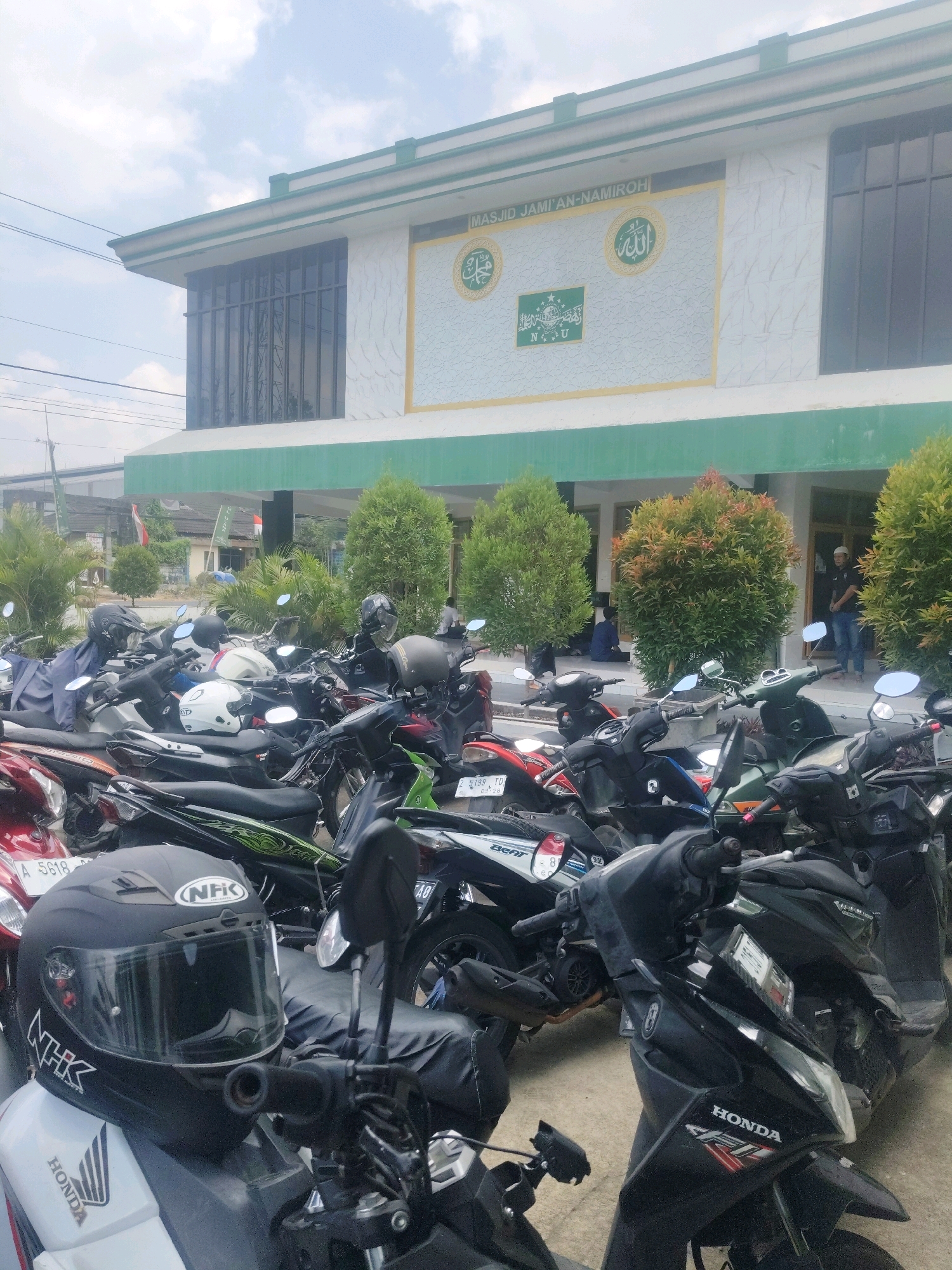 The image depicts the exterior of Masjid Jami'an-Namiroh in Kertasari, Indonesia, with a large number of parked motorcycles in the foreground and midground. The mosque has a distinctive facade with the name inscribed above decorative Islamic calligraphy and symbols. Several people are visible in the background near the entrance of the mosque, some sitting and others standing. The motorcycles are parked in a dense arrangement, suggesting a busy period. The environment appears to be outdoors during daylight, with some trees and vegetation surrounding the building. The overall scene captures a common sight in Indonesia, where motorcycles are a primary mode of transportation and often parked in large numbers outside public places like mosques.