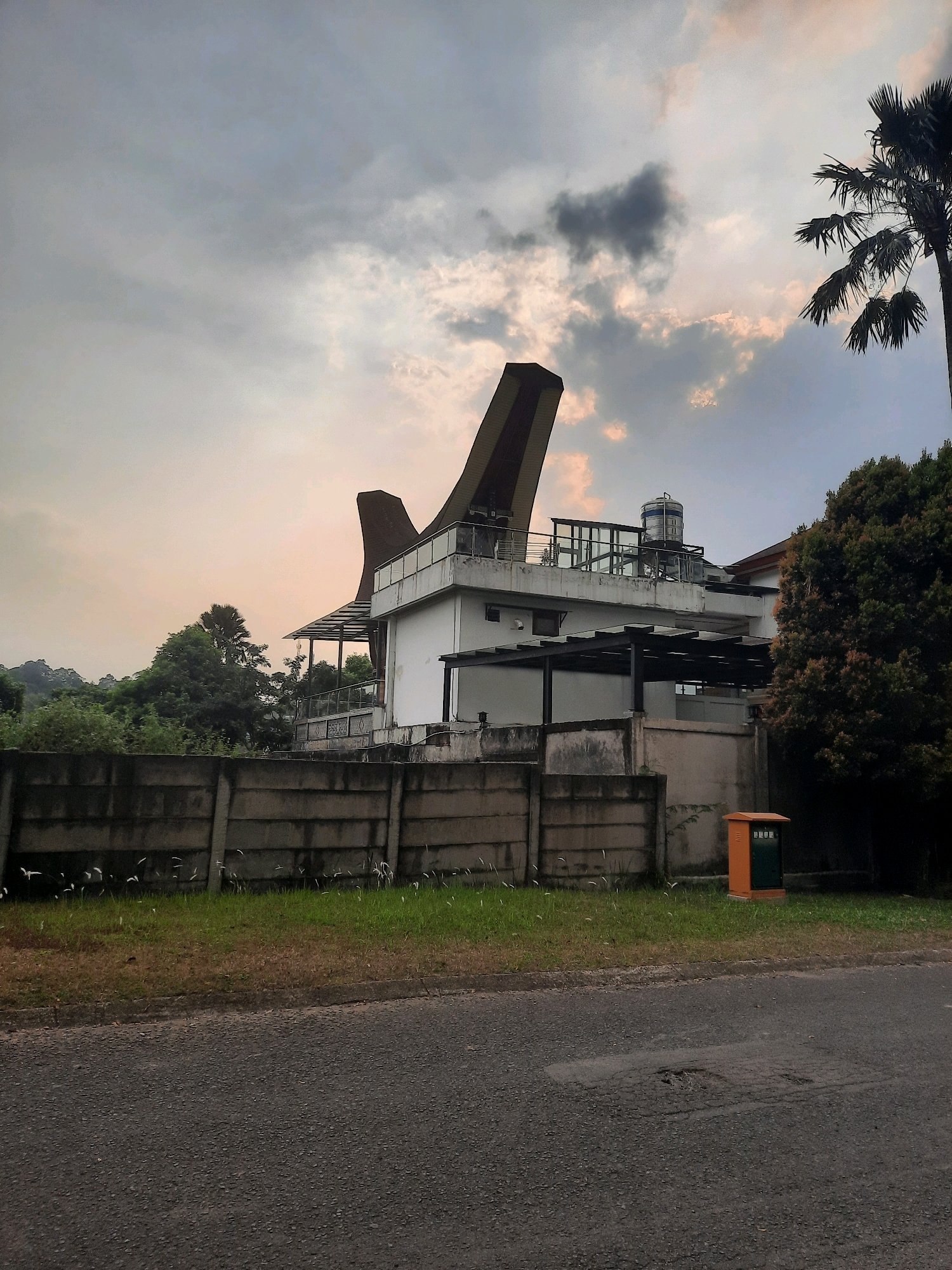 The image depicts a street view in Babakan Madang, Indonesia, with a distinctive modern house in the background. The house features a white facade and a unique, angled roof structure with dark brown accents. A balcony with glass railings runs along the upper level, and a water tank is visible on the rooftop. In front of the house, a concrete fence separates it from the street, with a strip of green grass and some tall, wispy plants along its base. To the right of the fence, an orange and green utility box stands on the grass. The street in the foreground is paved with asphalt and shows signs of wear, including a pothole. A tall palm tree with fanned fronds is visible on the right side of the frame, silhouetted against a cloudy sky. Lush green trees and vegetation form a backdrop to the house. The sky is overcast with patches of light suggesting either dawn or dusk.