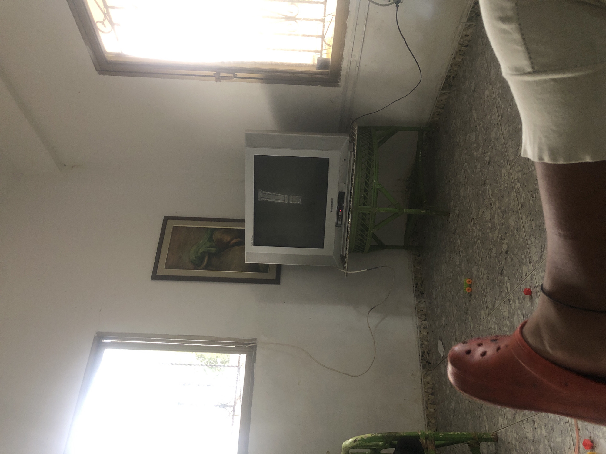 The image depicts an indoor scene, likely a living room, characterized by natural light streaming in from two windows. The walls are painted white and show signs of age and wear, with some marks and smudges. A framed painting, featuring a greenish subject, hangs on the wall between the windows.
Dominating the center of the room is a television set, appearing to be an older cathode-ray tube model, placed on a green, wire-mesh stand. The screen displays an exclamation mark, suggesting an error message or no signal. To the left, a portion of a green chair is visible.
The floor is tiled with a gray, speckled pattern. On the floor, in the foreground, a person's leg is visible, wearing an orange clog. Several small, colorful objects, possibly children's toys, are scattered on the floor.
The overall impression is one of a simple, somewhat cluttered living space, potentially in a residential setting in La Tagua, Venezuela, given the characteristic architecture and atmospheric lighting. The presence of the older television and the casual arrangement of objects suggest a lived-in environment.