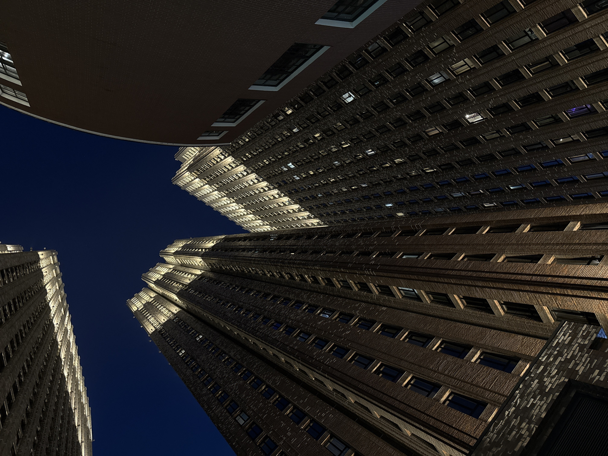 The image captures a low-angle view of multiple tall buildings against a dark blue sky, likely at night. The buildings are constructed of dark brick, and have many windows, some of which are illuminated. The architecture appears modern with some Art Deco influences. The location is potentially in Moscow, Russia, given the architectural styles present. There are no people visible in the scene. The focus is on the built environment and the perspective creates a dramatic and imposing effect.