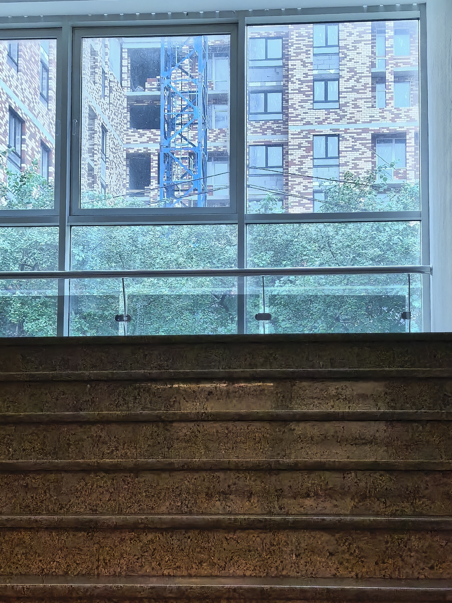 In an urban setting, a view through a large window reveals construction in progress. To the left, a brick building under construction is partially visible. In the center and to the right, a taller, unfinished brick building with multiple windows is the dominant feature. A bright blue construction crane is positioned in front of the right portion of the brick building. Below this upper window, a lower window looks out onto dense green foliage, suggesting trees or shrubs. A glass and metal railing separates the interior space from the view through the lower window. In the foreground, a close-up view of rough, textured, layered stairs made of a dark, weathered material fills the bottom portion of the frame. The overall lighting is subdued, creating a somewhat muted color palette.