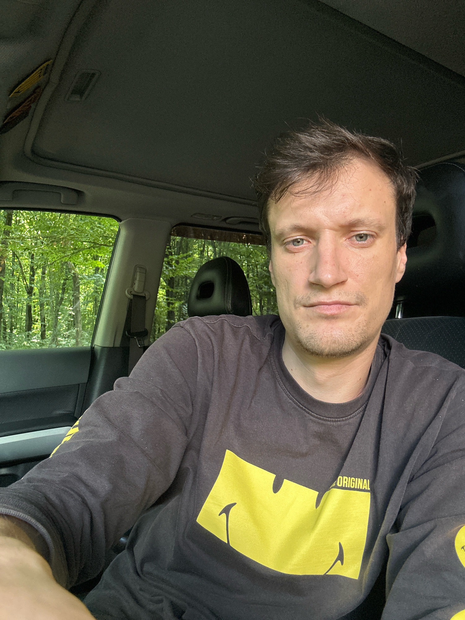 The image is a selfie taken from inside a vehicle. The subject is a man with brown hair and light-colored eyes, wearing a dark long-sleeved shirt with a yellow smiley face graphic. He is sitting in the driver's seat, and the interior of the car, including the ceiling with a sunroof, is visible. Outside the car, through the windows, a dense forest with green foliage can be seen. The scene appears to be set in a natural, possibly rural, environment. The location of Shatava, Ukraine is not determinable from the image content.