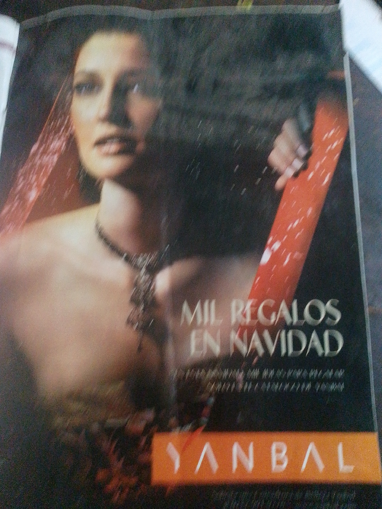 The image shows a close-up of a magazine or advertisement featuring a woman with a veiled head and a necklace. Text overlays the image, with "MIL REGALOS EN NAVIDAD" (A THOUSAND GIFTS FOR CHRISTMAS) prominently displayed. Below this, the word "YANBAL" is visible in large letters on an orange background. The context suggests a promotional item for the beauty brand Yanbal, possibly related to Christmas sales. The location in Santa Ana, Venezuela, is not directly visible or indicated within the image itself, but could be inferred from the context of where this advertisement was found. The image quality is low, with blurriness and graininess, making precise details difficult to discern.