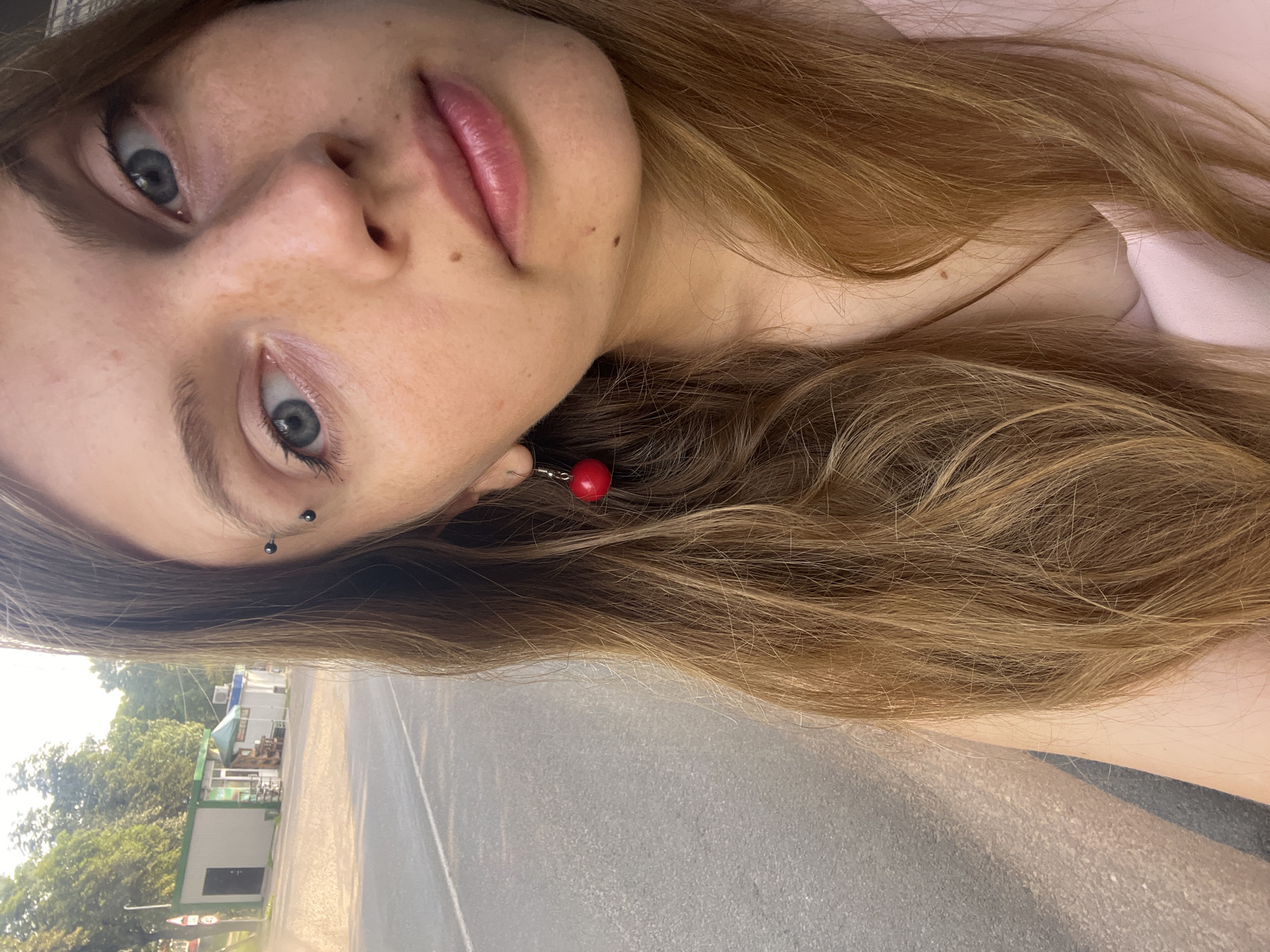 This is a selfie of a young woman with long, wavy blonde hair and blue eyes. She is wearing a light pink top and has a black eyebrow piercing and a red cherry-shaped earring. Her face is turned towards the camera, and she has a light smattering of freckles across her nose and cheeks. Her lips are slightly parted, and she appears to be smiling subtly.
In the background, there is a paved road and a small, light-colored building with a green roof, possibly a kiosk or small shop. There are also trees and a lamppost visible. The lighting suggests it is daytime, possibly late afternoon due to the warm sunlight casting shadows.
The location is identified as Senitski Sel'ski Savet, Belarus. This rural administrative unit is located southwest of Minsk, the capital of Belarus. The scene depicts a roadside environment that is typical of such a location, with elements of infrastructure (road, building) and natural surroundings (trees).