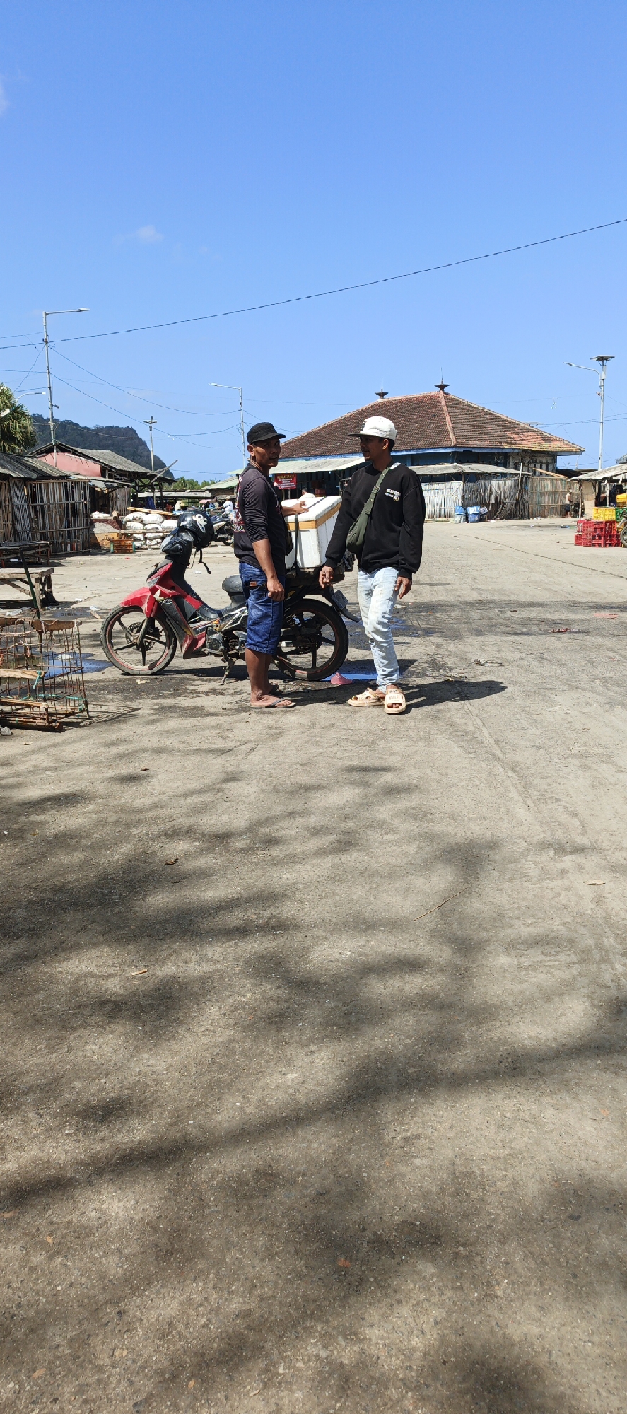 In Puger, Indonesia, two men stand on a dusty road, facing each other in conversation. One man, wearing a black long-sleeved shirt and a black cap, has his left hand extended, appearing to point at or gesture towards a white cooler box mounted on the back of a red motorcycle. He wears blue shorts and flip-flops. The other man, in a black sweatshirt and a white cap, has his arms down by his sides and wears light-colored jeans and sandals. He holds a green bag slung over his right shoulder. The motorcycle is red and black, with a black helmet resting on its seat. To the left of the motorcycle is a wire cage with some wooden material. In the background, there are several wooden stalls and buildings with tiled roofs, suggesting a market or a fishing village environment. A bright blue sky with scattered clouds is visible above. The ground is paved with a rough, grey asphalt surface with shadows cast across it, indicating a sunny day.