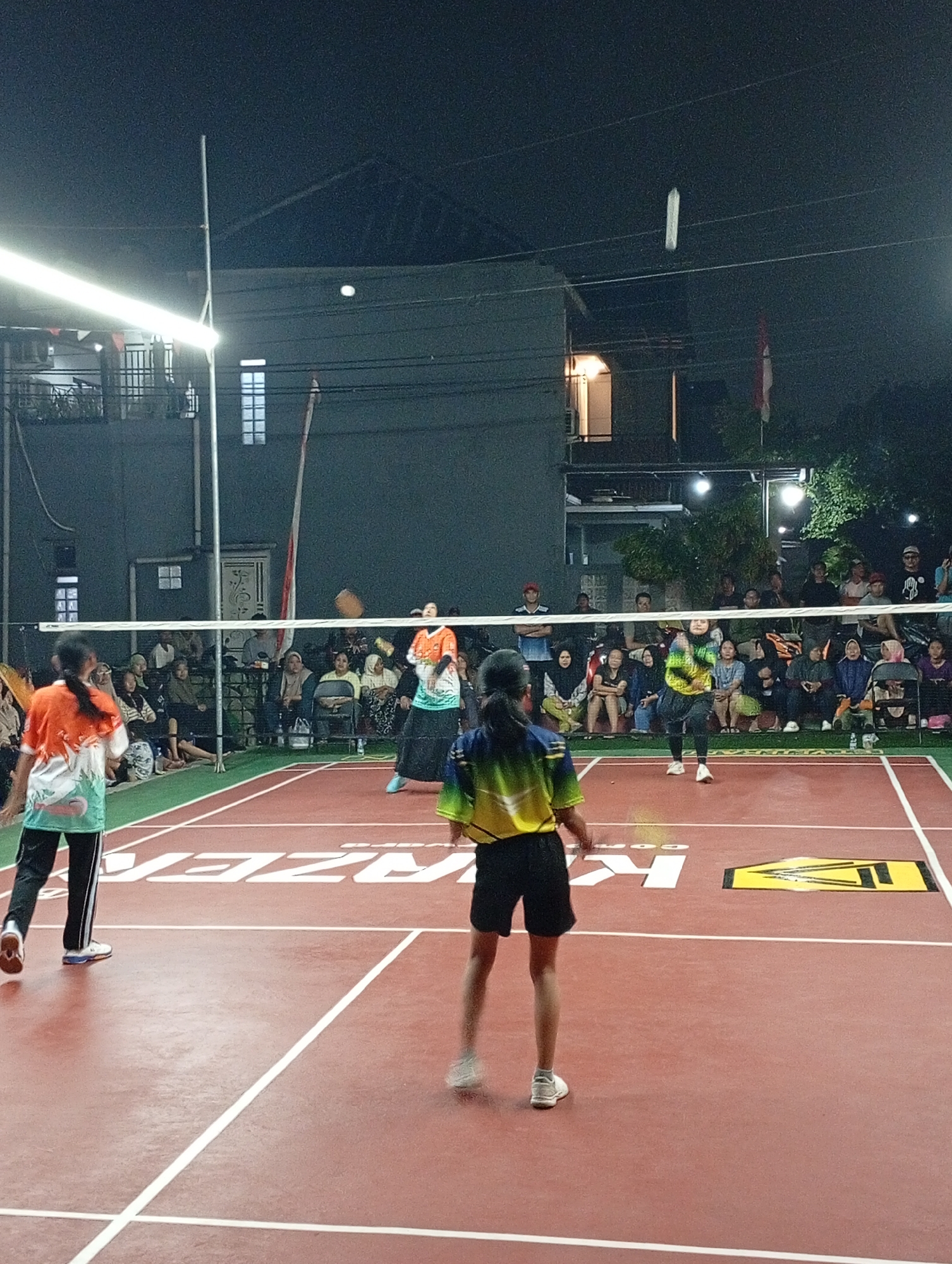 The image depicts an outdoor badminton match at night, possibly in Caringin, Indonesia, given the presence of an Indonesian flag in the background. A net divides the court, with four young female players actively engaged in the game. Two players are on one side of the net and two on the other. One player in the foreground on the left wears an orange and white shirt and black pants. On the right side of the net, a player in a yellow and green shirt and black shorts is captured mid-action, appearing to serve or hit the shuttlecock. The court is a reddish-brown color with white lines marking the boundaries. Spectators are seated on benches and on the ground behind the court, watching the match. The background shows buildings and trees, illuminated by artificial lights, creating a somewhat dimly lit atmosphere typical of an evening sporting event.