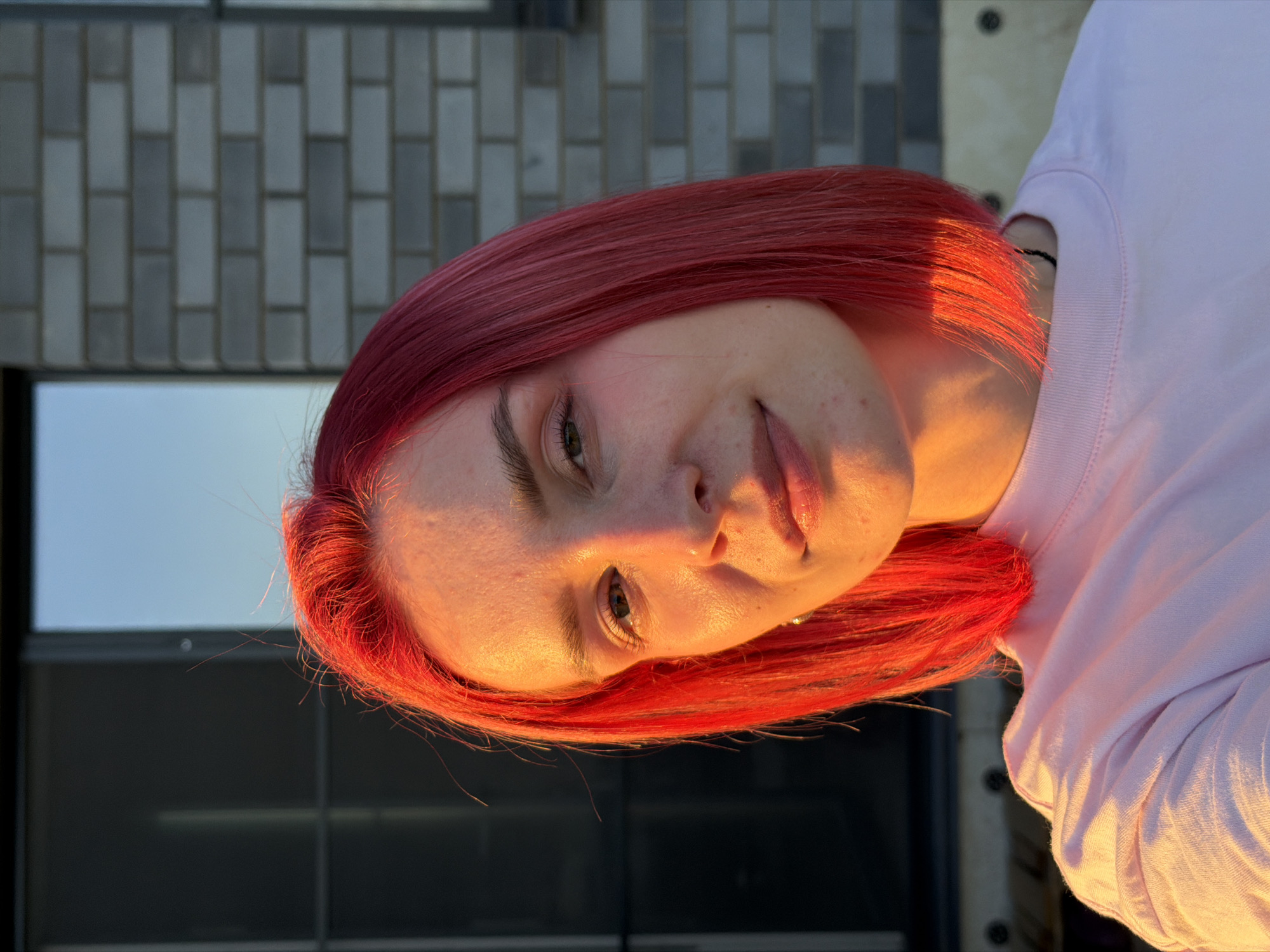 A young woman with short, bright red hair is pictured outdoors during golden hour, with the warm sunlight casting a glow on her face and light pink t-shirt. She is looking towards the camera with a slight smile. In the background, a modern building with grey brickwork and dark windows is visible. The overall lighting suggests it is late afternoon or early evening. The location is identified as being in the city of Chaikovsky, Russia.