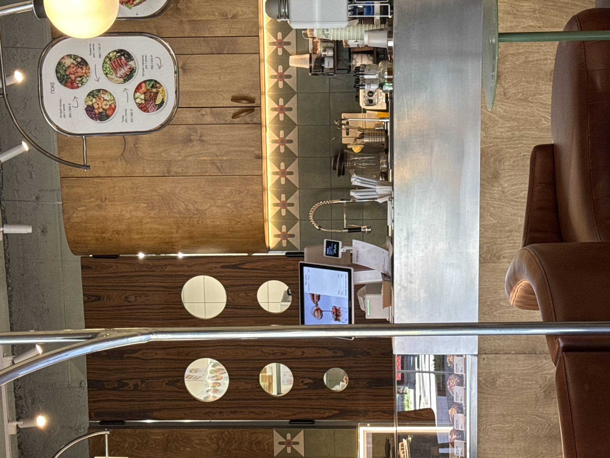 This image captures the interior of a modern and inviting cafe or restaurant, likely located in Sochi, Russia, as indicated by the Russian text on the menu. The scene is devoid of people, suggesting a quiet moment during the day.
On the left side, a prominent oval-shaped menu board hangs, displaying various "ПОКЕ" (Poke) bowls. Each option is presented with an appealing photograph and includes its name in Russian, such as "Острый лосось" (Spicy Salmon), "Тунец" (Tuna), "Креветка" (Shrimp), and "Острая говядина" (Spicy Beef). The descriptions mention "рис / кинов" (rice / quinoa) as base options, with prices listed in Rubles (P), for example, 850/900 P for Spicy Salmon. Bright light fixtures illuminate this section.
Extending across the middle and right of the frame is a sleek stainless steel counter, serving as a preparation area. It is equipped with various coffee-making components, blenders, glasses, and utensils, all neatly arranged. Behind the counter, a distinct tiled wall or floor section features a repeating geometric pattern of light-colored crosses on a dark green background, enhanced by warm strip lighting. Above this, a segment of light-colored wooden paneling adds to the decor.
In the foreground, a dark wooden panel, possibly a partition or part of a serving station, features several circular decorative elements. Some of these are reflective mirrors, while others appear to be simple cream-colored tiles. A tablet displaying an image of someone pouring a drink is visible on this panel, likely used for ordering or display. To the far right, a comfortable brown leather armchair provides seating, positioned next to a small, circular glass-topped table with a green stand. The overall design harmoniously blends natural wood tones with modern stainless steel and subtle geometric patterns, creating a contemporary and warm atmosphere.