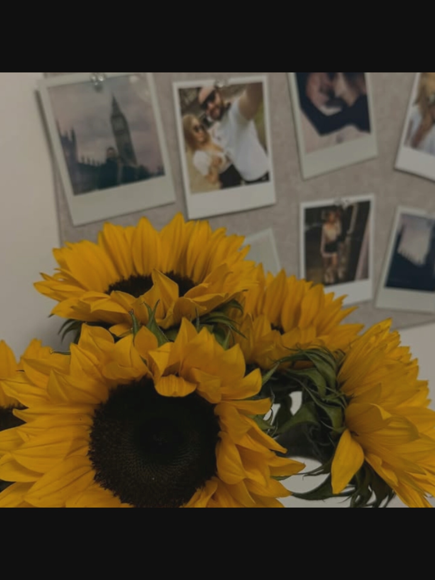 The image shows a bouquet of sunflowers in the foreground, with several framed photos attached to a wall in the background. The photos appear to be instant prints and depict various scenes including a cityscape that may be London (featuring a clock tower resembling Big Ben), a couple taking a selfie, another couple, and individuals in different settings. The sunflowers are bright yellow with dark centers and green stems. The overall lighting is soft, suggesting an indoor environment. The backdrop of the photos is a light-colored wall or board. There is no visible indication or context connecting this image to the city of Bucharest, Romania.