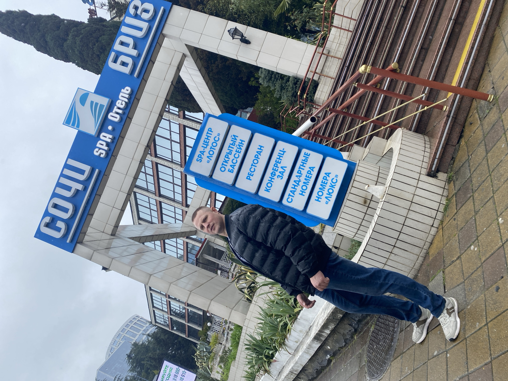 A full shot of a man standing on a sidewalk, with a large sign for the Sochi Breeze SPA hotel in the background. The sign is blue and white with Russian text. The man is wearing a black jacket, blue jeans, and grey sneakers. He is looking towards the camera. The hotel is a large, modern building with many windows. There are stairs and railings visible in the background. The weather appears overcast. There is a sign with Cyrillic text offering a service. The sidewalk is made of paving stones.