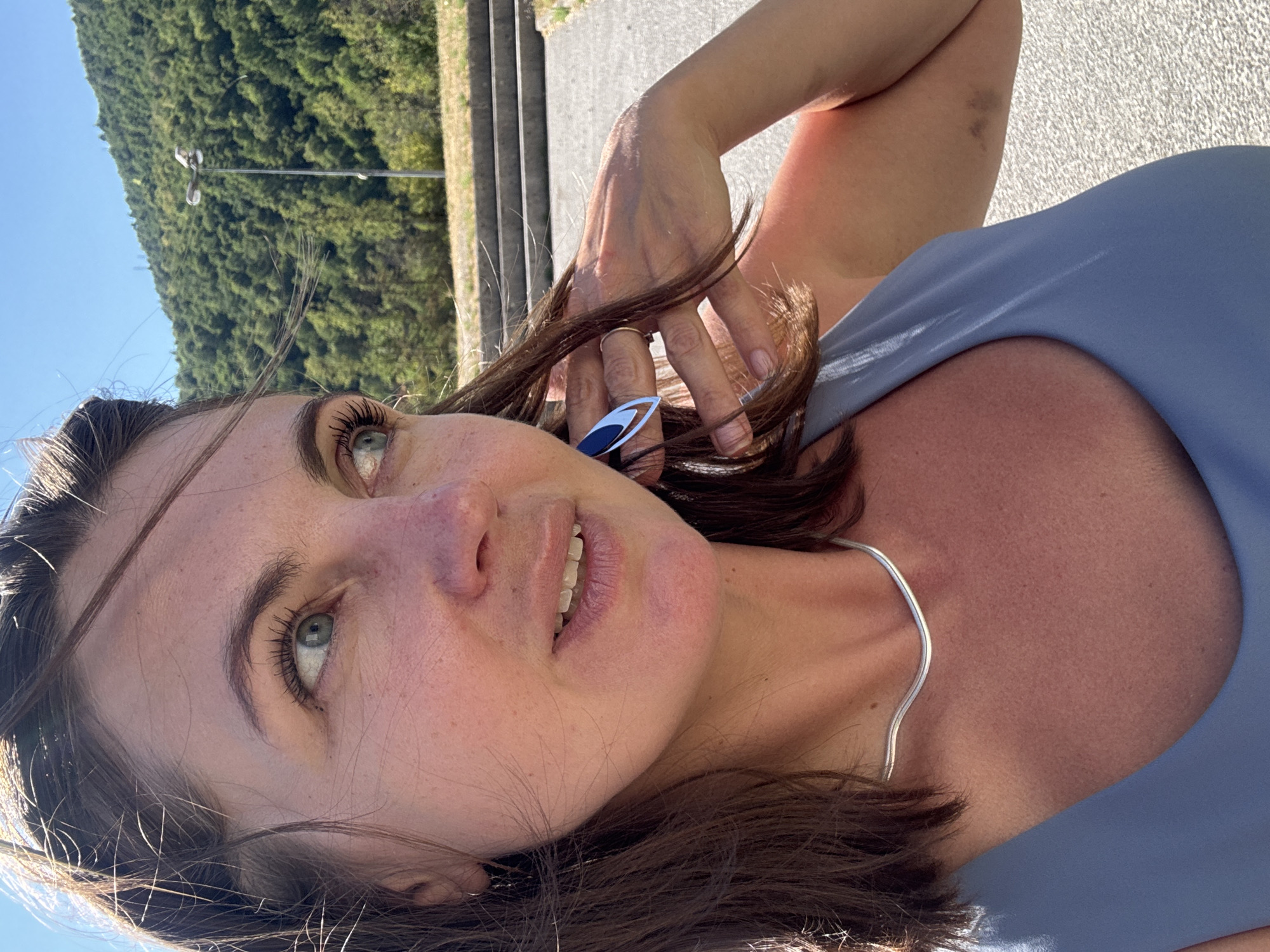 A medium shot captures a woman in a blue top, seen from the chest up, as she poses for a selfie outdoors on a sunny day. Her brown hair, styled with a side part, is windswept, with strands framing her face. She has fair skin with light freckles and green eyes. She's wearing a silver necklace that rests on her collarbone and a ring on her right index finger. Her right hand is raised, touching her hair near her ear. Her left ear has a silver earring with a dark blue element. The background is a blurred image of a hillside covered in dense green trees under a clear blue sky. In the mid-ground, there are concrete stairs and a lamppost. The ground in the foreground is a paved surface. The scene appears to be set in a natural or semi-natural outdoor area, potentially a park or roadside overlook in Saint-Claude, France, given the prevalence of lush, forested hillsides in the region.