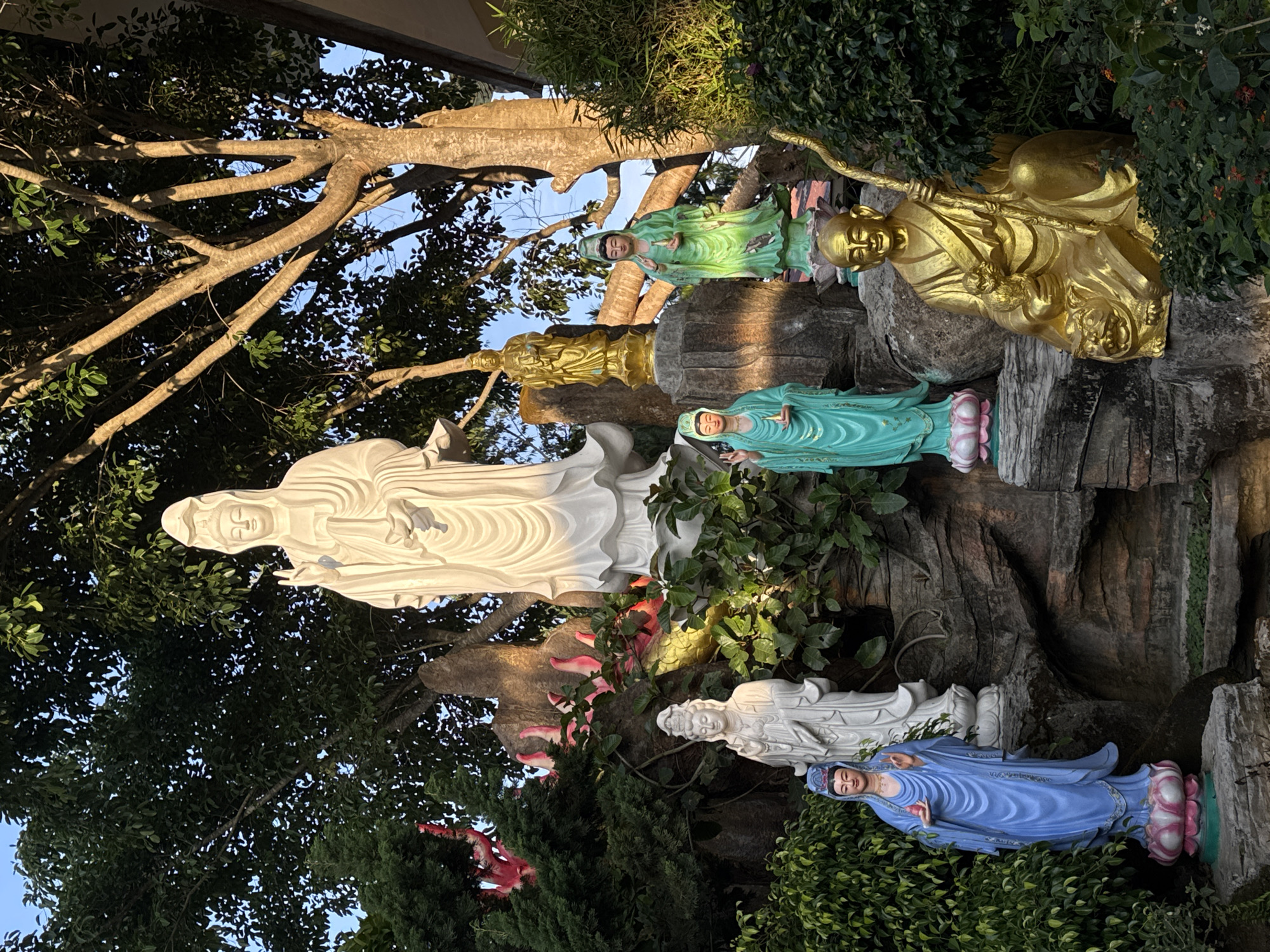 The photograph captures a statue garden in Lâm Viên, Đà Lạt, Vietnam. A tall, white statue of a female deity dominates the left side of the image, set against a backdrop of dense green foliage and tree branches. Adjacent to it and slightly higher up are statues of a golden monk figure, and green robed figure. Further down the slope are white and blue deities. They are placed on stone structures resembling a rocky hillside. The scene is bathed in natural light, with the tree branches creating shadows and dappled light.