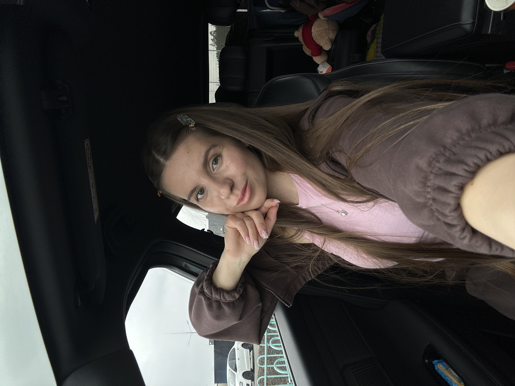 A young woman with long brown hair is seated in the driver's seat of a vehicle. She is wearing a pink shirt and a brown jacket. A hair clip is visible in her hair. The car is parked near a building, potentially in Vtoraya Alekseevka, Russia, although the location is not definitively confirmed within the image itself. Outside the window, there is a white vehicle and what appears to be a metal railing. In the back seat of the car, a stuffed animal is present.