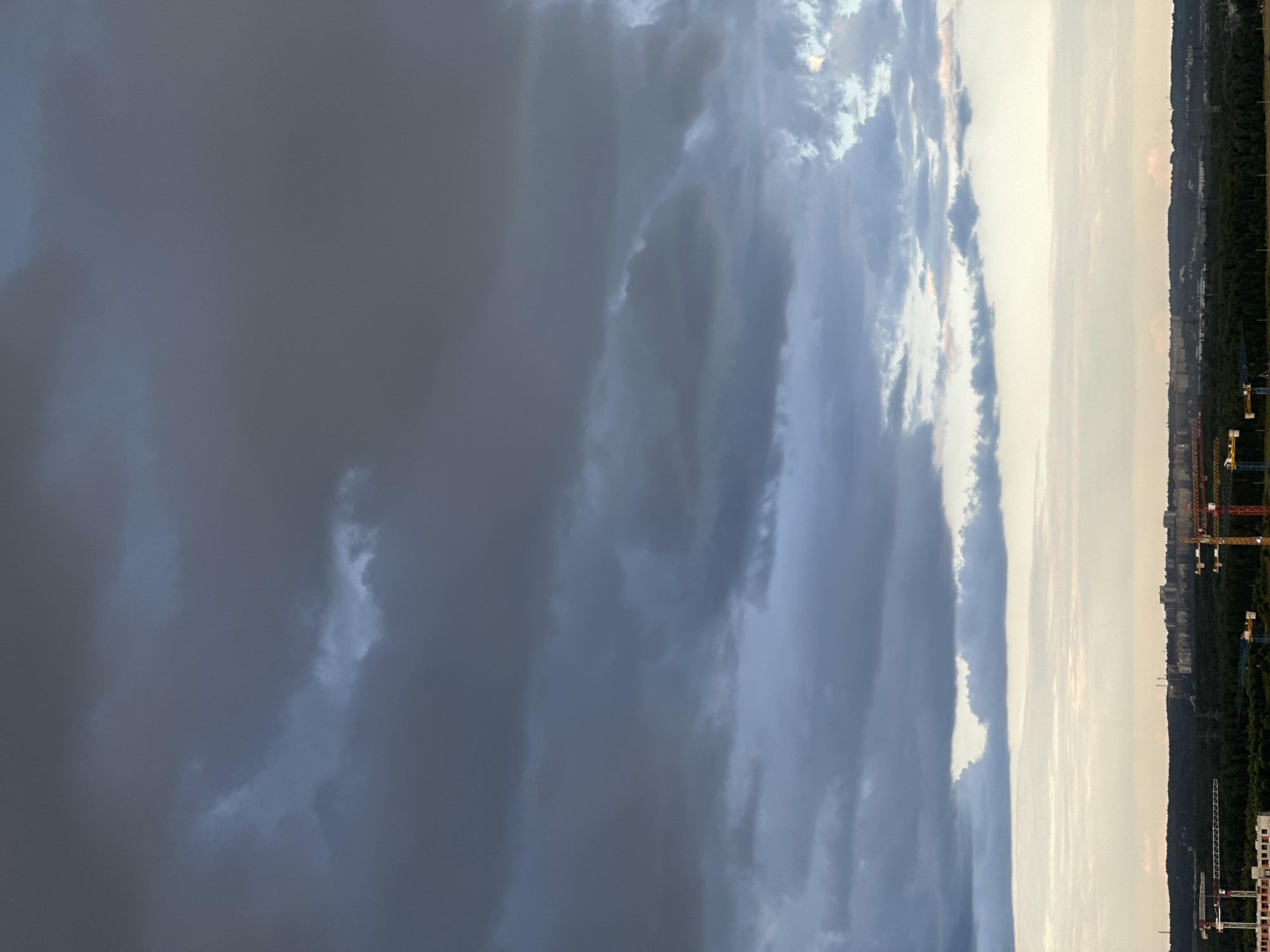 The image depicts a panoramic view of a city skyline under a dramatic sky filled with dark, heavy clouds. The lower portion of the image shows a densely forested area with several construction cranes and buildings visible on the horizon. The city of Dnp Ryzhiki, Russia, is identified as the location. The scene appears to be during the late afternoon or early evening, with the clouds suggesting an impending storm or the aftermath of one. The sky is a mix of dark grays and blues, with some lighter patches where the sun may be breaking through. The cityscape in the distance is a collection of apartment buildings and industrial structures. The foreground features construction sites with multiple cranes, indicating ongoing development. No people are visible in the frame. The overall impression is one of urban expansion against a backdrop of imposing natural elements.