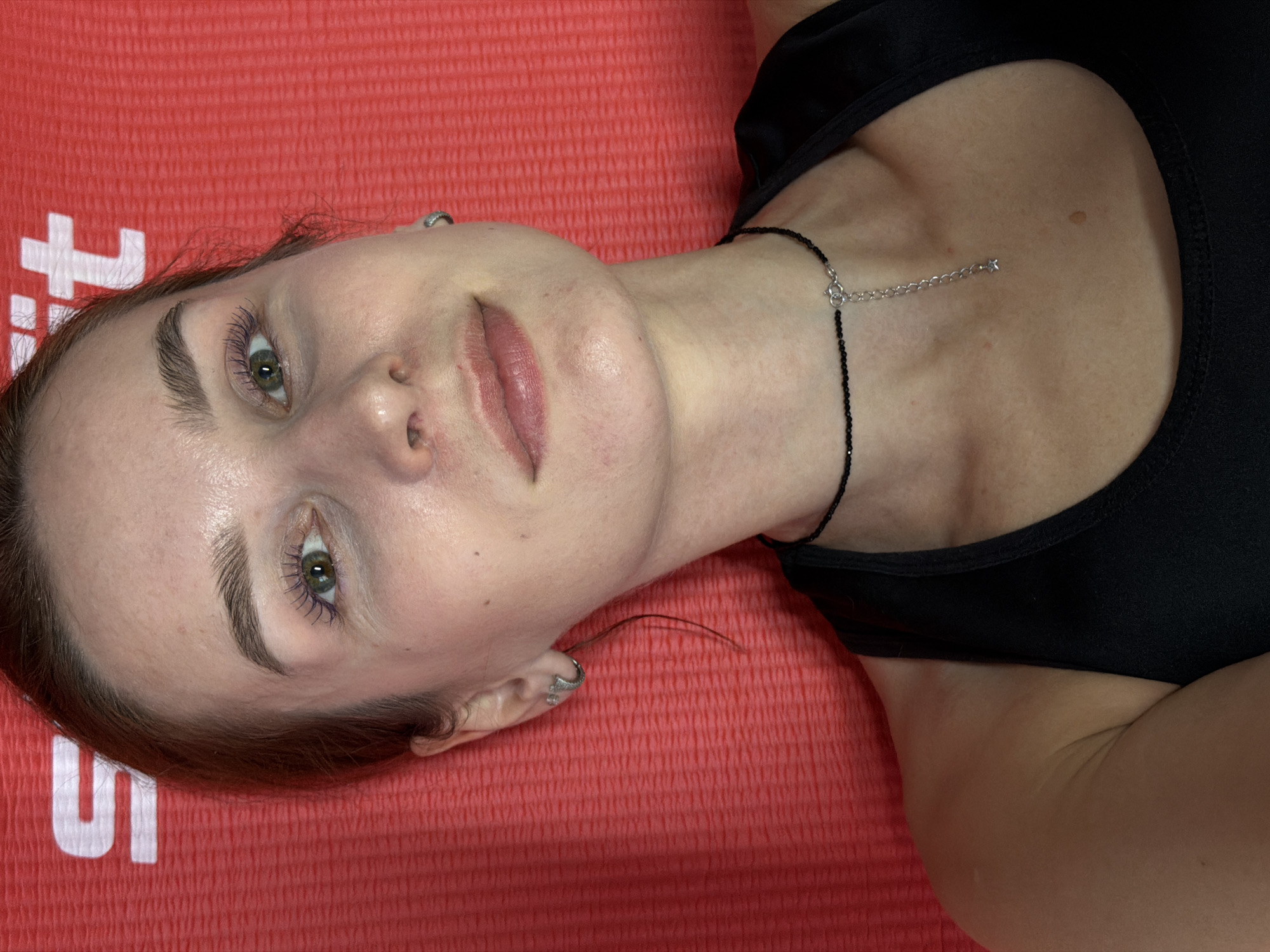 A medium shot depicts a woman laying on a textured orange surface. She is wearing a black tank top and a black beaded choker with a silver chain extender. The woman has fair skin, green eyes, and light brown hair pulled back. Her eyelashes appear to be coated in a purple mascara. She is wearing silver earrings. The image's orientation suggests the photo was taken from a close, elevated angle. The orange surface underneath the woman appears to be a yoga mat, displaying sections of white lettering at the bottom left. The presence of the yoga mat suggests this image was taken in Chaikovsky in a physical fitness club, gym, yoga studio, dance studio, or home fitness area.