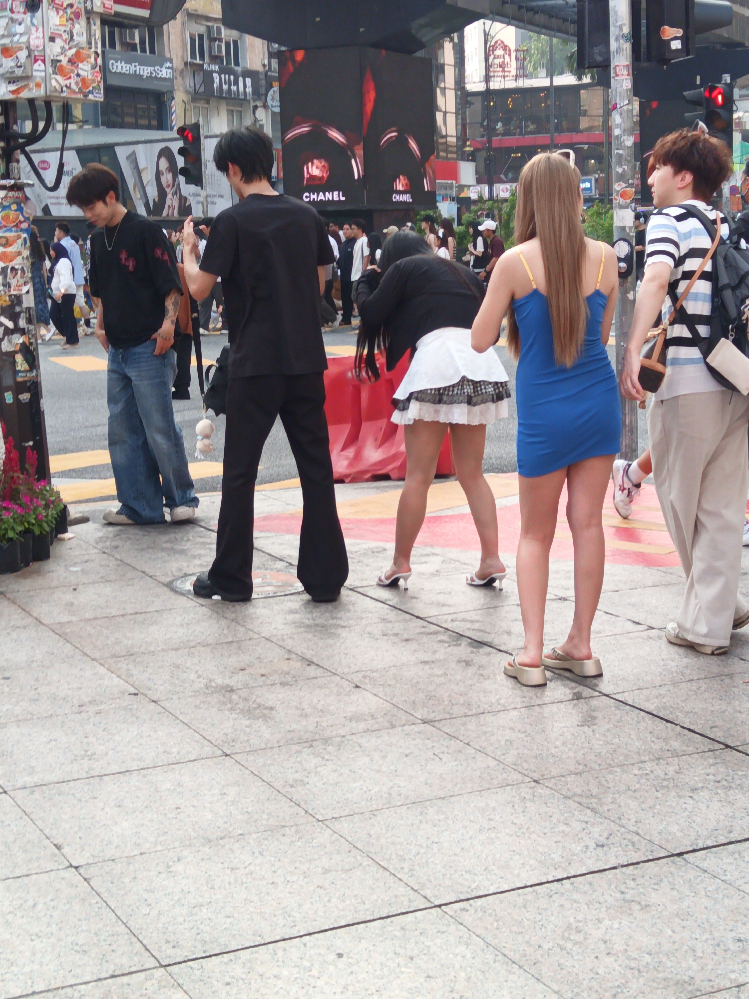 The image depicts a street scene in Kuala Lumpur, Malaysia, likely in the Bukit Bintang area given the prominent advertisements and urban environment. Four individuals are visible walking on a paved sidewalk.
On the left, a man wearing a black t-shirt and blue jeans is walking towards the viewer, with his hands in his pockets. Next to him, another man in a black t-shirt and black flared pants walks away from the viewer, carrying a black bag over his shoulder.
In the center, a woman in a white mini skirt and a black top walks away from the viewer, her back is turned to the camera. Beside her, a woman in a vibrant blue dress with yellow straps also walks away from the viewer.
In the background, there are buildings with various advertisements, including a large screen displaying the "CHANEL" logo. Traffic lights and other urban elements suggest a busy city setting. The sidewalk is made of large, grey tiles. The overall impression is a snapshot of daily life in a bustling urban environment.