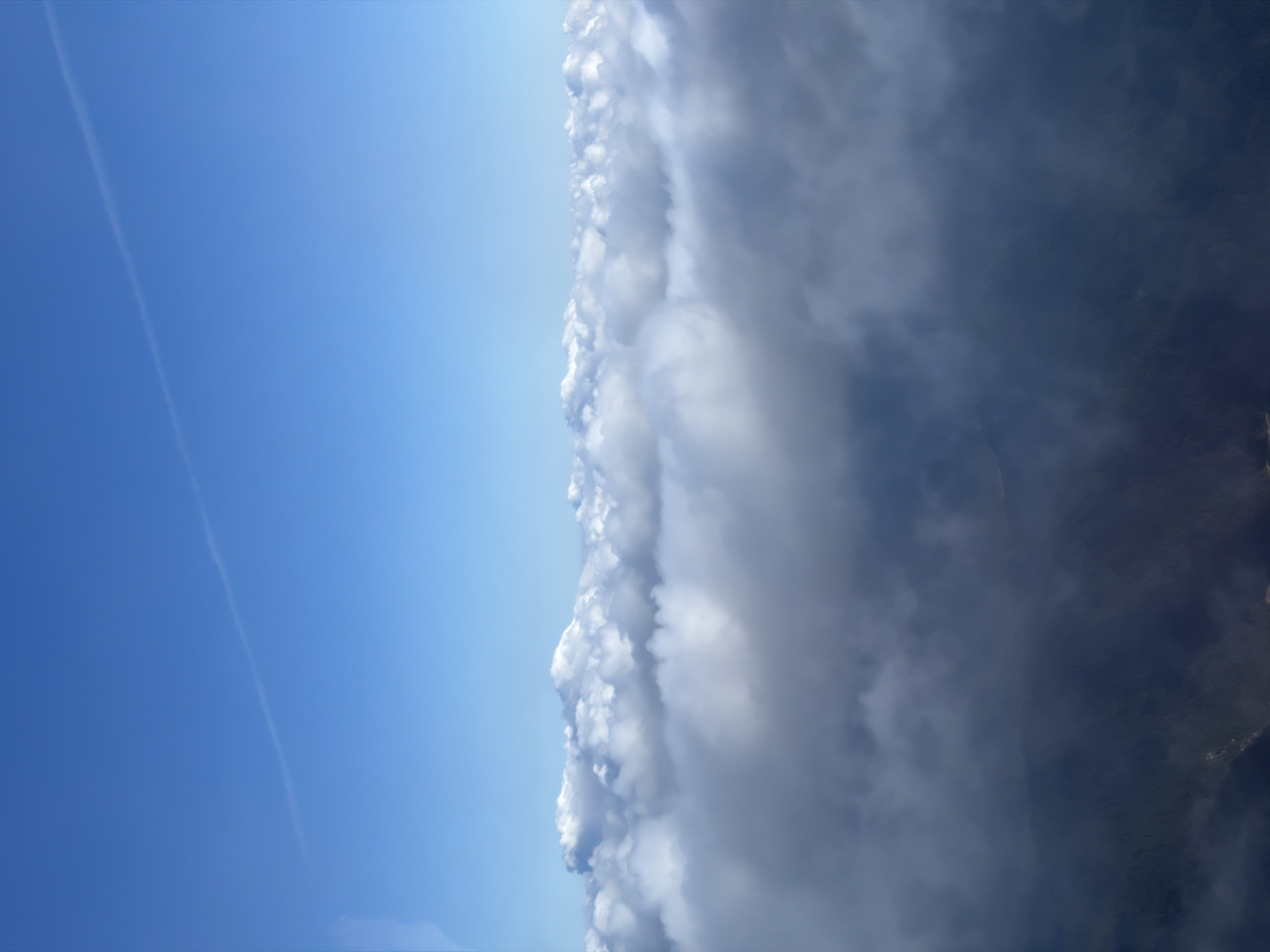 This aerial photograph depicts a clear blue sky with a thin contrail stretching diagonally from the upper left to the upper right. Below the sky, a thick layer of fluffy white clouds forms a distinct horizon line. The lower portion of the image is obscured by darker, more diffuse clouds, through which the faint outlines of a mountainous landscape can be discerned. It is impossible to determine the specific location within Grainau, Germany, or identify any people, subjects, objects, or actions in this image due to the natural, atmospheric subject matter.