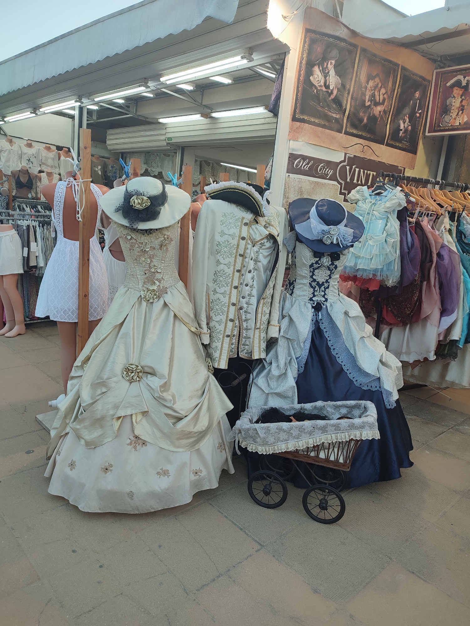 This image shows a retail stall selling vintage-style clothing, likely in Nesebar, Bulgaria, as indicated by the presence of "Old City Vintage" signage. Two mannequins are prominently displayed, one wearing a cream-colored, elaborate gown with floral embellishments and a wide-brimmed hat, and the other in a dark blue and white gown with a matching hat. Behind them, a man's ornate, embroidered jacket is displayed on a mannequin. A rack of colorful, tiered dresses is visible in the background, along with framed prints of people in historical attire. A small, antique-style baby carriage with lace trim sits in front of the blue and white dress mannequin. The stall appears to be outdoors, with a light-colored awning overhead, and the ground is paved with stone tiles.