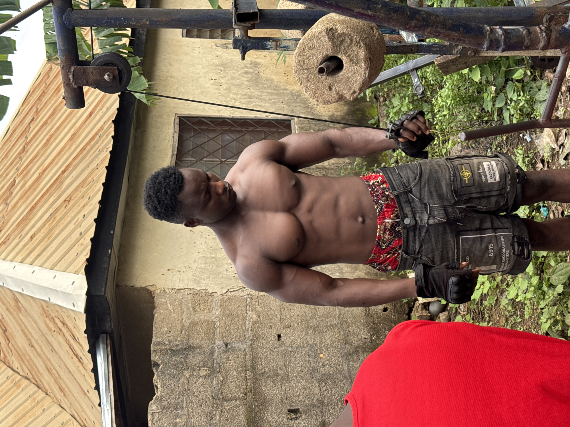 In Akwanga, Nigeria, a shirtless Black man with a muscular physique stands outdoors, looking down. He is wearing black fingerless gloves and grey cargo shorts with a red and gold patterned waistband. The man is positioned in front of a building with a corrugated metal roof, a stone wall, and two windows. To his right, a makeshift weightlifting apparatus is visible, featuring a stone weight and a pulley system. Green foliage surrounds the area. In the foreground to the left, a portion of another person wearing a red shirt is visible.