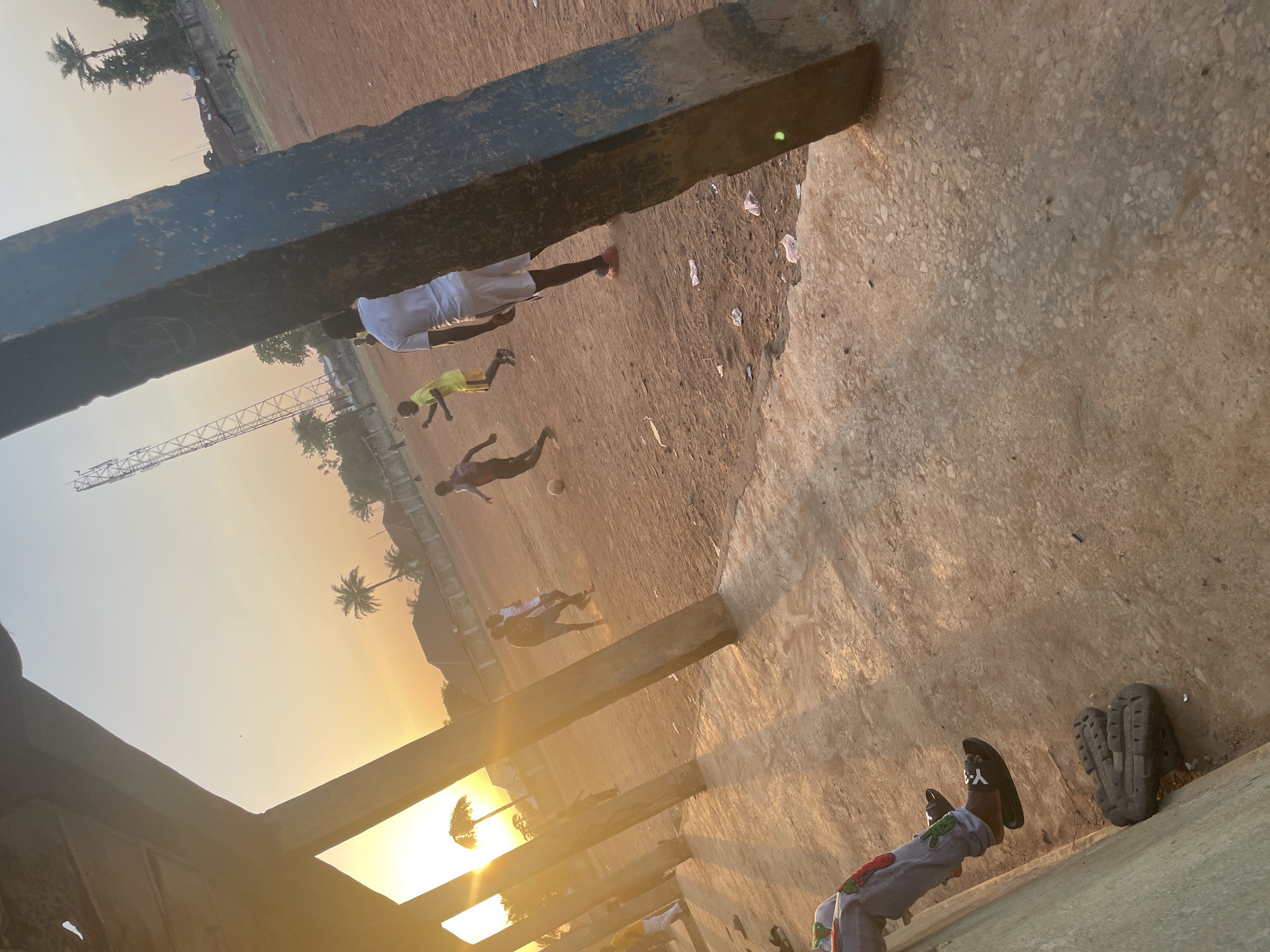 A partially obscured, downward-angled view in Akwanga, Nigeria shows several people playing soccer on a dirt field during sunset. The lower right corner displays a concrete structure, with two pairs of footwear including sandals. The foreground partially obscures the view of the field, where at least four individuals are engaged in soccer. Beyond the field are trees and buildings silhouetted against the sun. An overhead tower is also visible.