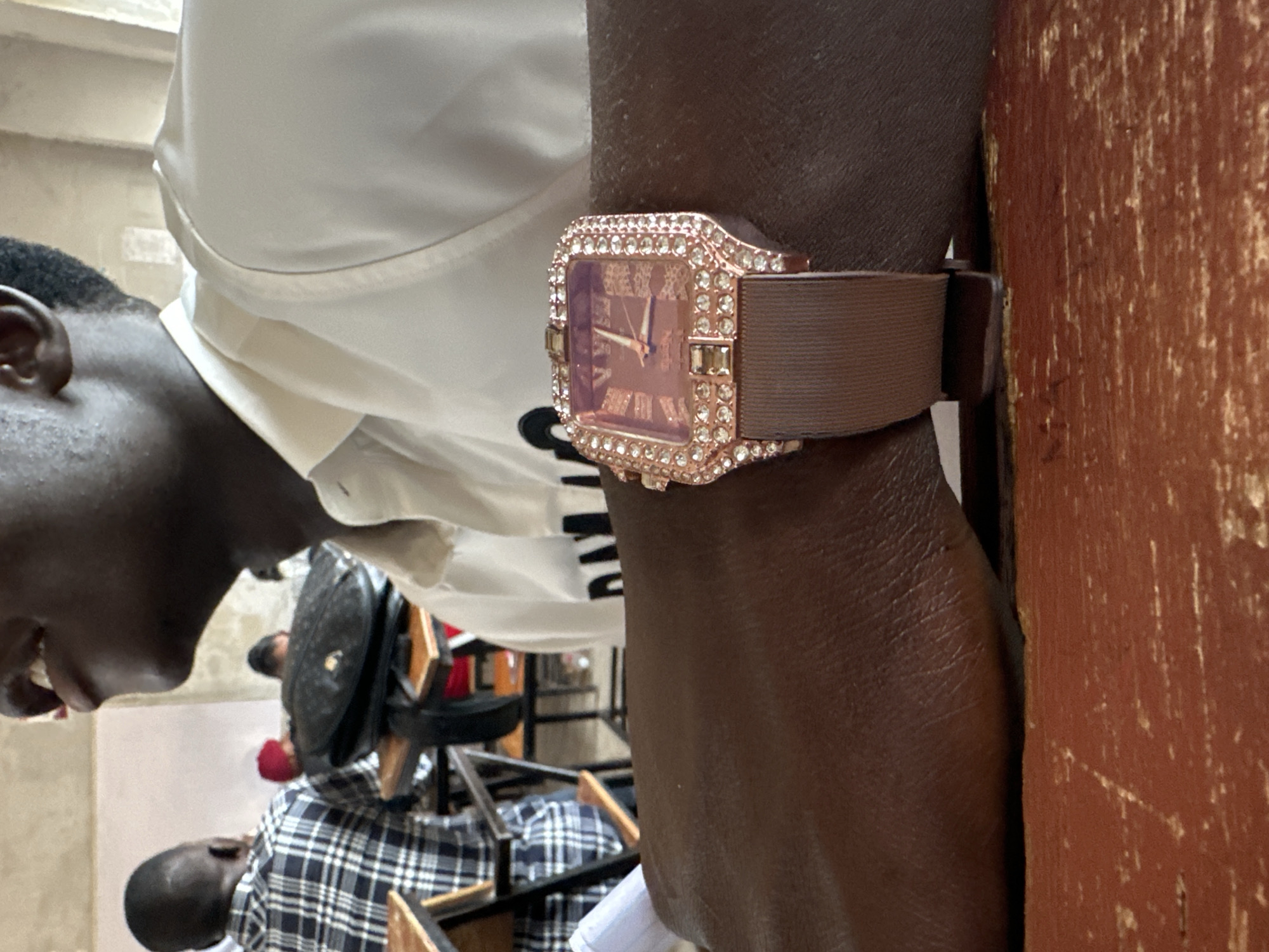 A medium shot depicts a person's arm wearing a rose gold-toned watch with a brown strap. The watch face is adorned with rhinestones. The person is wearing a white shirt. A second person wearing a plaid shirt is visible in the background, along with chairs and other people in the distance. A painted wall is visible on the right side of the frame. The location is Jos, Nigeria.