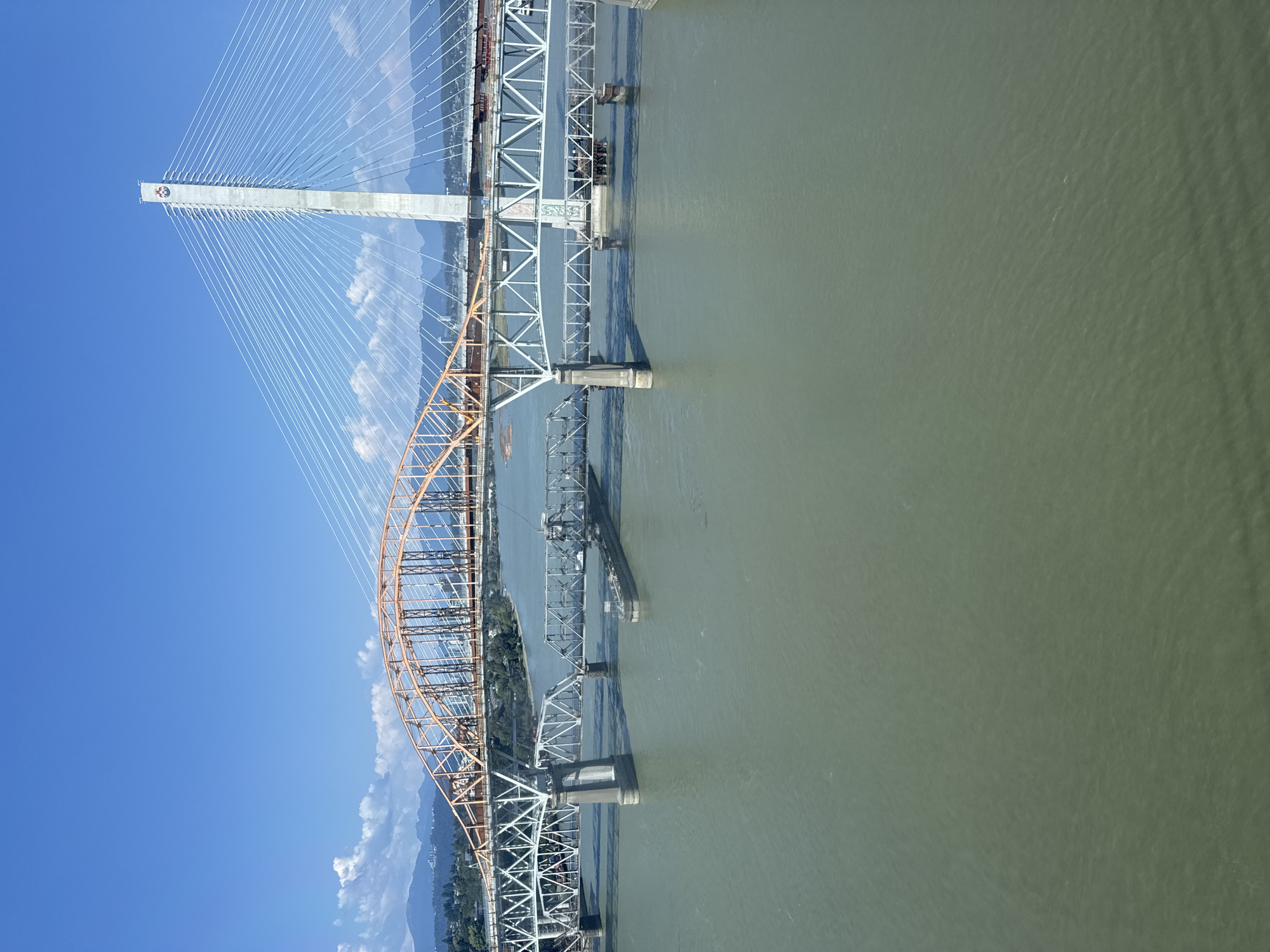 The image shows an aerial view of the Fraser River in New Westminster, Canada, with multiple bridges spanning across it. The foreground is dominated by the murky green water of the river. In the middle ground, there are two distinct bridges. The bridge closer to the foreground is a lower, steel truss bridge with concrete piers. A long barge is visible beneath this bridge. Behind it, a larger, higher bridge with an orange arch structure is visible. Further in the background, a modern cable-stayed bridge with a tall white pylon is seen, with numerous cables extending from the pylon to the bridge deck. The background shows a distant cityscape and mountains under a clear blue sky with scattered white clouds. The overall scene depicts a busy riverine transport and infrastructure hub.