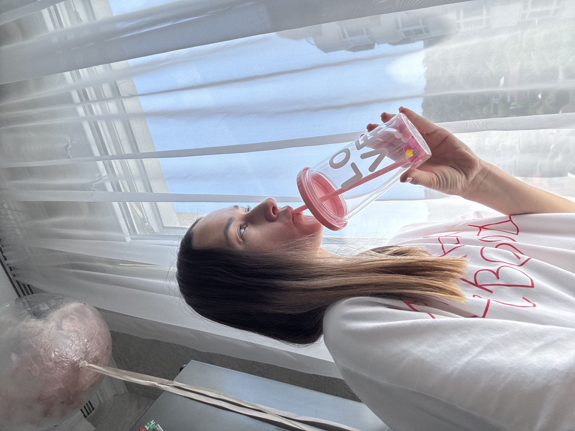 The image shows a close-up, rotated perspective of a woman indoors, illuminated by natural light from a window. She is depicted from the chest up, with long, straight brown hair falling over her shoulder. Her head is tilted upwards, and she is looking towards the upper right of the frame (if rotated upright). She is actively drinking from a clear plastic tumbler with a vibrant pink lid and straw. The word "LOVE" is clearly visible, spelled vertically in white letters on the side of the tumbler, which also contains small pink, flower-shaped decorative elements near the lid. Her right hand, adorned with light red nail polish, holds the tumbler.
She is wearing a white t-shirt featuring red outlined letters, partially visible across her chest and shoulder. The background is dominated by a large window covered with white horizontal blinds or sheer curtains, allowing bright daylight to filter in. Beyond the window, a bright blue sky and faint outlines of structures, possibly buildings, are discernible, suggesting a daytime scene.
In the bottom left foreground, a large, translucent, pinkish-white spherical object, which appears to be a decorative balloon or a large, fluffy confection on a stick, is partially visible. The scene captures a casual, relaxed moment during the day in an indoor setting, consistent with the provided location context of Sochi, Russia.