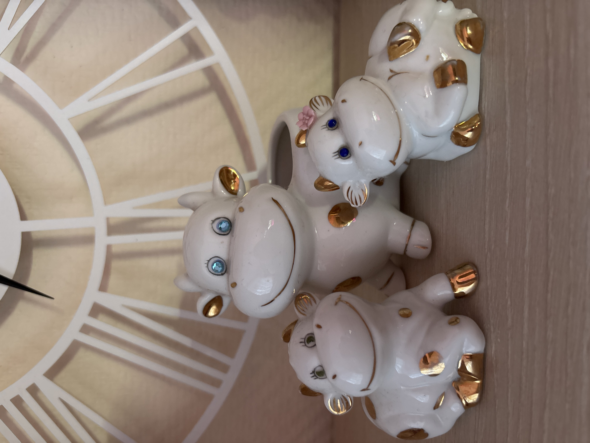 The image displays an indoor scene in Русский Вожой, Russia, featuring three glossy white ceramic cow figurines with gold-colored accents, positioned on a light-colored wooden surface. In the background, a light beige wall is partially obscured by a white, open-work wall clock featuring Roman numerals and a black minute hand pointing towards the upper right quadrant.
The leftmost figurine is a seated calf with green rhinestone eyes, painted black eyelashes, and a smiling mouth. It has irregular gold-colored spots on its body and gold hooves.
The central figurine is a standing cow with blue rhinestone eyes, painted black eyelashes, and a smiling mouth. It features gold-colored spots and hooves, and an open cavity is visible on its back, suggesting a planter or holder function.
The rightmost figurine is a reclining calf with blue rhinestone eyes, painted black eyelashes, and a smiling mouth. It exhibits gold-colored spots and hooves, and a small pink flower embellishes its head near the left ear.
The wooden surface beneath the figurines shows a subtle grain pattern. The wall clock's white frame and numerals contrast with the light beige wall behind it.
