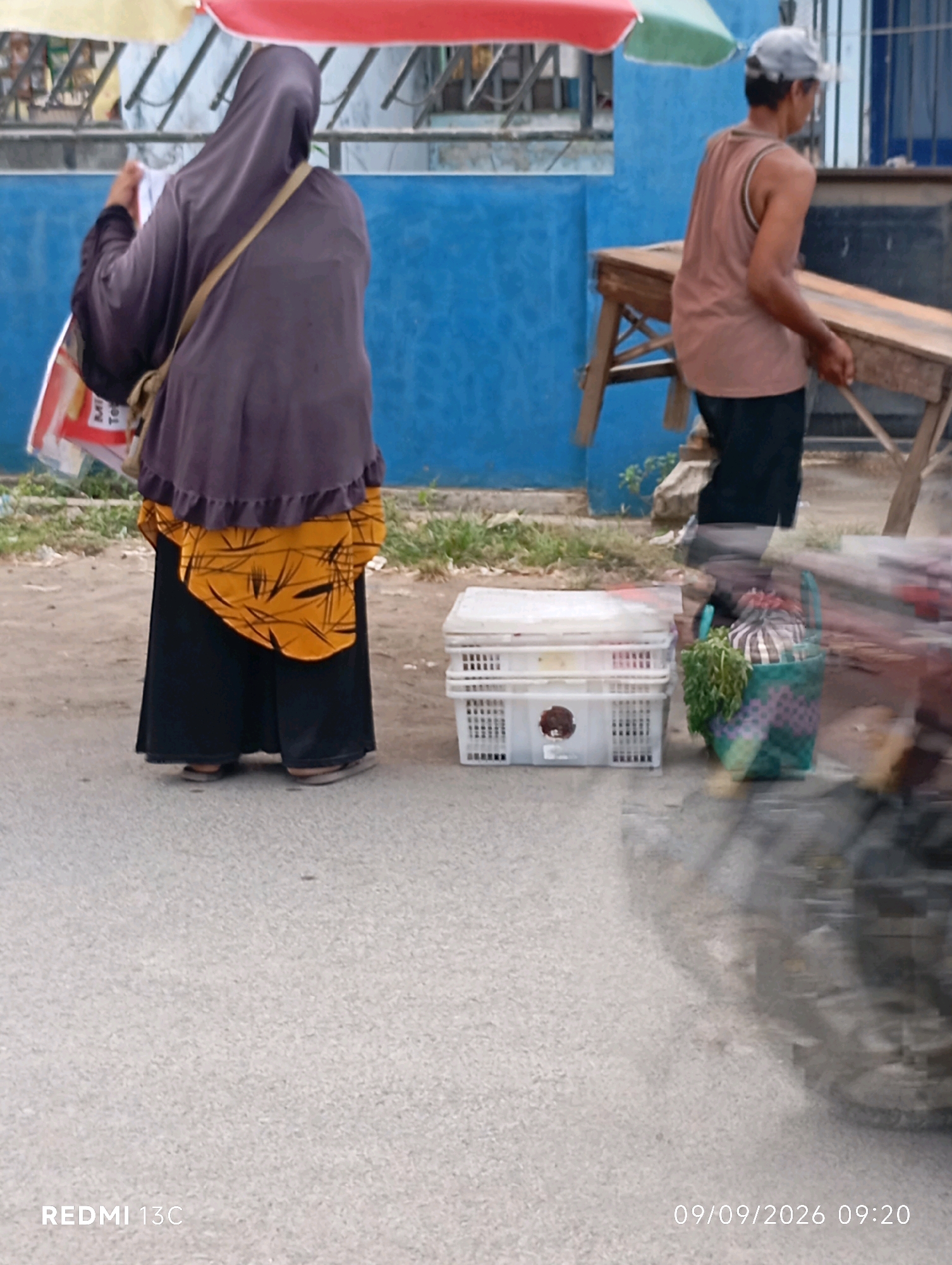 In Wuluhan, Indonesia, two individuals are observed on a paved road, with a blue building forming the backdrop. On the left, a person wearing a dark purple hijab and a yellow patterned blouse over a long black skirt stands with their back to the camera. They are carrying a stack of papers in their right hand and a light-colored bag with a beige strap is slung over their left shoulder. To the right, a person wearing a light brown tank top and dark trousers, with a grey cap on their head, is near a wooden table. In front of this person, there are two white plastic crates, one stacked on top of the other. A green woven basket containing leafy green vegetables and a striped object is placed beside the crates. The scene appears to be outdoors, with a blurred motion effect suggesting movement, possibly from a passing vehicle. The timestamp "09/09/2026 09:20" is visible in the lower right corner of the image.