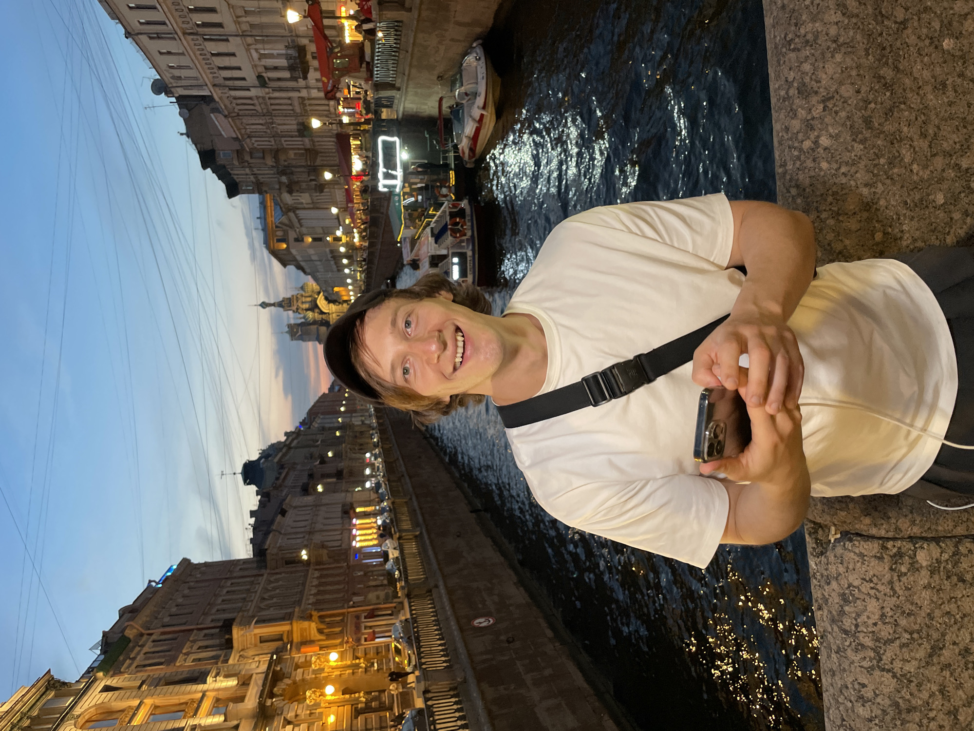 An outdoor, eye-level, medium-shot photograph shows a young, Caucasian man smiling while holding a mobile phone and standing on a stone bridge. The man is wearing a white t-shirt, dark pants, and a black baseball cap, backwards. He has light brown hair, and is looking directly into the camera. A black strap bag is slung across his chest. The background is a canal lined with ornate, multi-story buildings that are illuminated with yellow lights. Several boats are on the canal, and the sky above is a pale blue with hints of orange from a setting sun. This scene is located in St. Petersburg, Russia, on a bridge over the Griboyedov Canal, with the Church of the Savior on Spilled Blood visible in the distance. The time of day is dusk, and the water in the canal reflects the lights from the buildings and the sky.