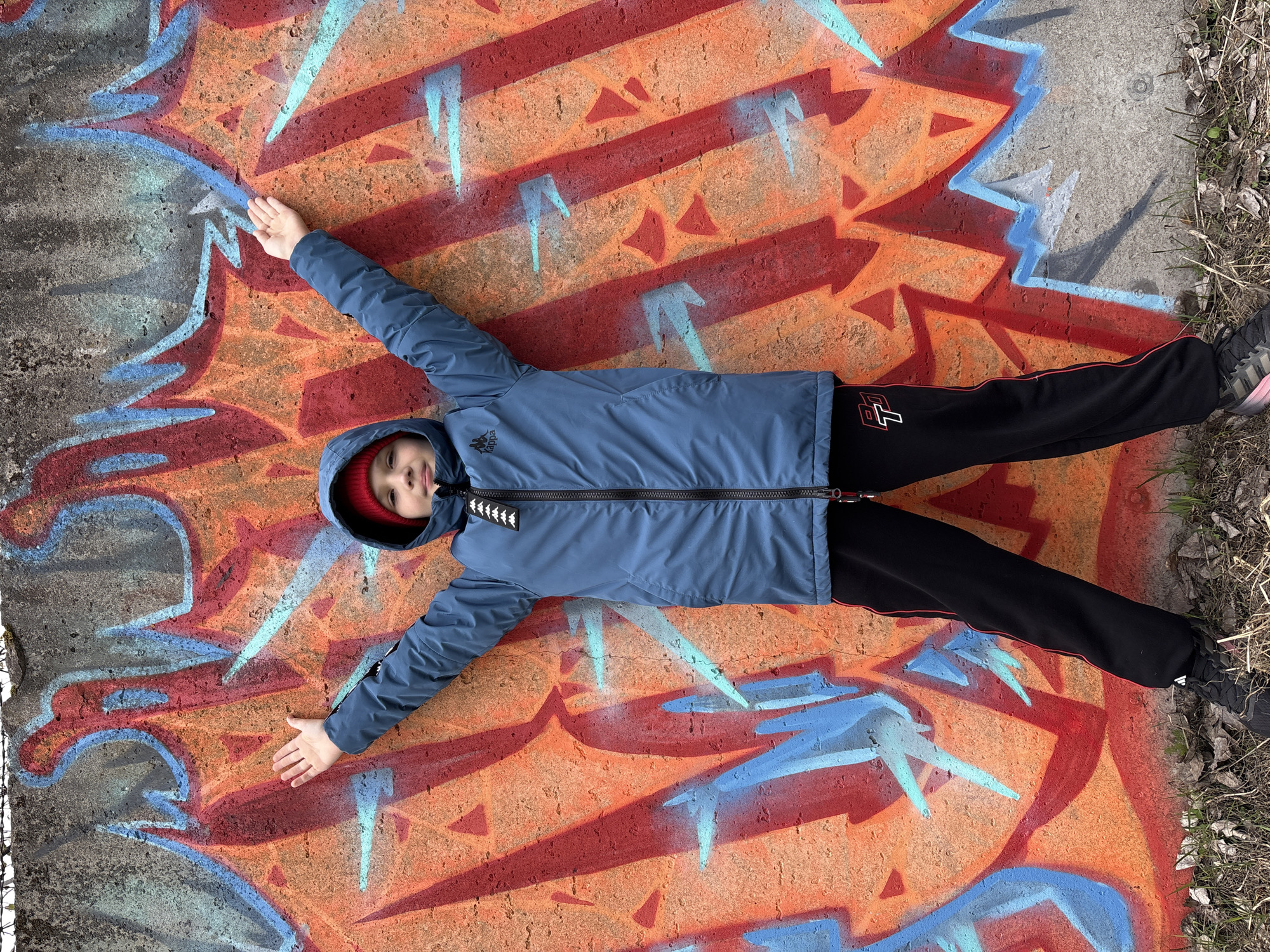 A boy is lying in a star shape on a concrete surface with graffiti in Chaikovsky, Russia. The graffiti is primarily orange, red, and blue, and depicts arrows and jagged lines. The boy is wearing a blue jacket, black pants with a red stripe, and a red hat. He is lying flat on his back with his arms outstretched. The surface is slightly uneven. Vegetation is visible to the right of the frame.