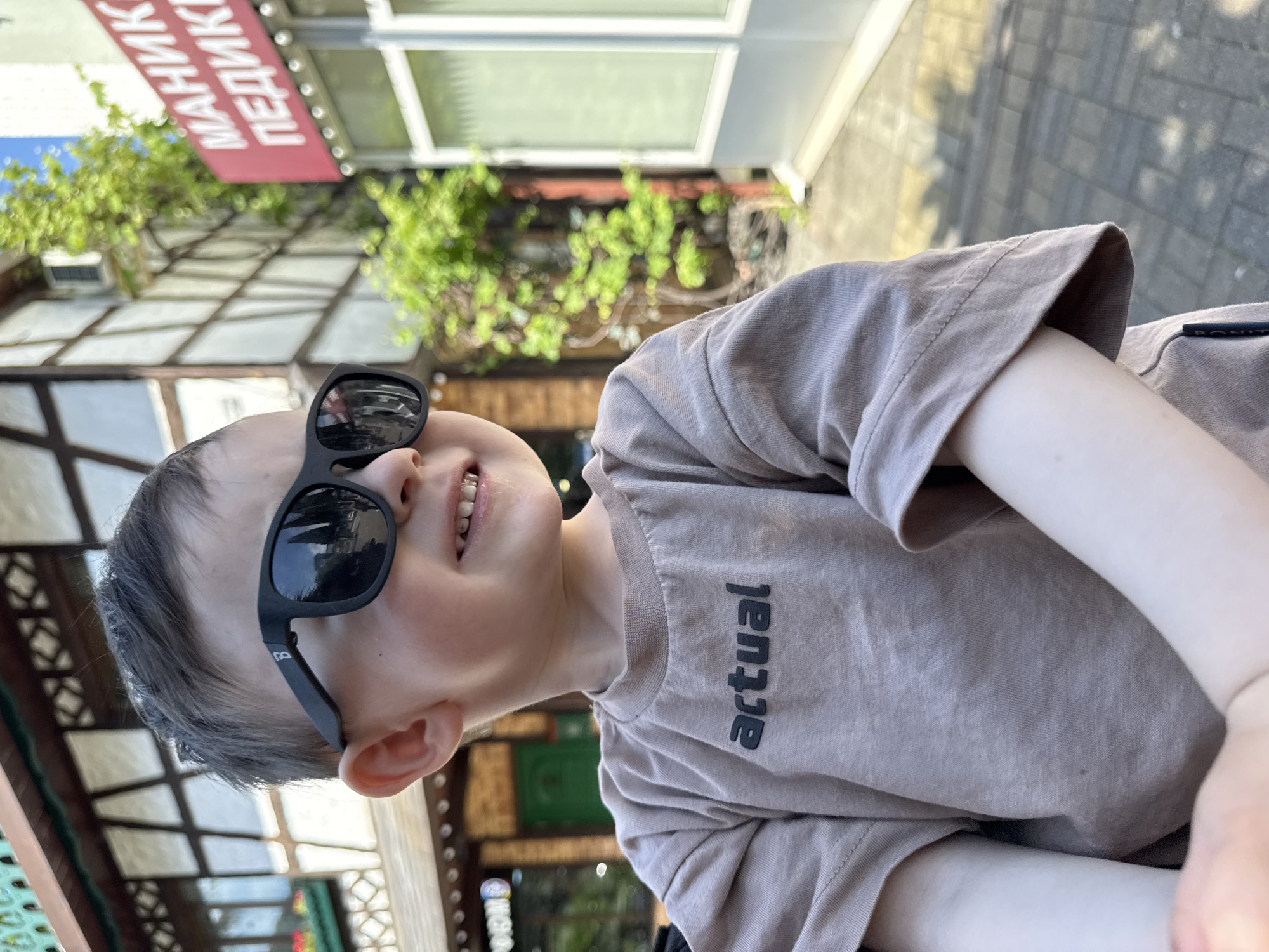 A young child wearing sunglasses and a beige shirt with the word "actual" on it is the primary subject of the image. The child is smiling and appears to be outdoors, possibly sitting down. The background includes elements of a building with a sign that reads "МАНИКЮР ПЕДИКЮР", suggesting a manicure and pedicure salon, and foliage. The architecture of the building and the signage indicate that the location is likely in a city where Russian is spoken. Given the context of previous prompts, the location is likely Sochi, Russia. The image has been rotated.