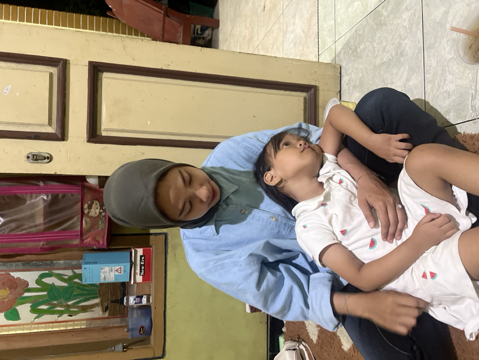 An adult female and a young girl are seated on a patterned brown and white rug in an indoor setting in Gabus, Indonesia. The adult female, wearing a dark olive-green hijab, a light blue long-sleeved collared shirt, and dark trousers, is positioned with her legs bent. Her left arm is around the young girl, with her left hand resting on the girl's lower torso. The adult's gaze is directed downwards. The young girl, dressed in a white short-sleeved collared dress patterned with small red and green watermelons, is leaning her head against the adult female's left arm, looking slightly upwards and to the right.
The background on the left side features a yellowish wall with a brown-framed window or wall art depicting an orange and green floral design. Below this, a blue "OPPO" brand box, a white "New-Era" box, a small blue container, and a clear bottle with a black cap (possibly hand sanitizer) are stacked. A pink transparent hanging organizer with a Hello Kitty design is visible on the wall. A white bag rests on the rug to the adult's left.
On the right side of the image, a light-colored wooden door with brown trim and a visible doorknob plate is partially open, revealing an outdoor area. This exterior area includes a terracotta-colored plastic or wooden chair and a light-colored brick wall. The floor beyond the rug consists of light-colored square tiles. A plastic cup with a straw, containing a beverage, is partially visible on the tiled floor in the bottom right corner. The scene is illuminated by artificial indoor lighting.