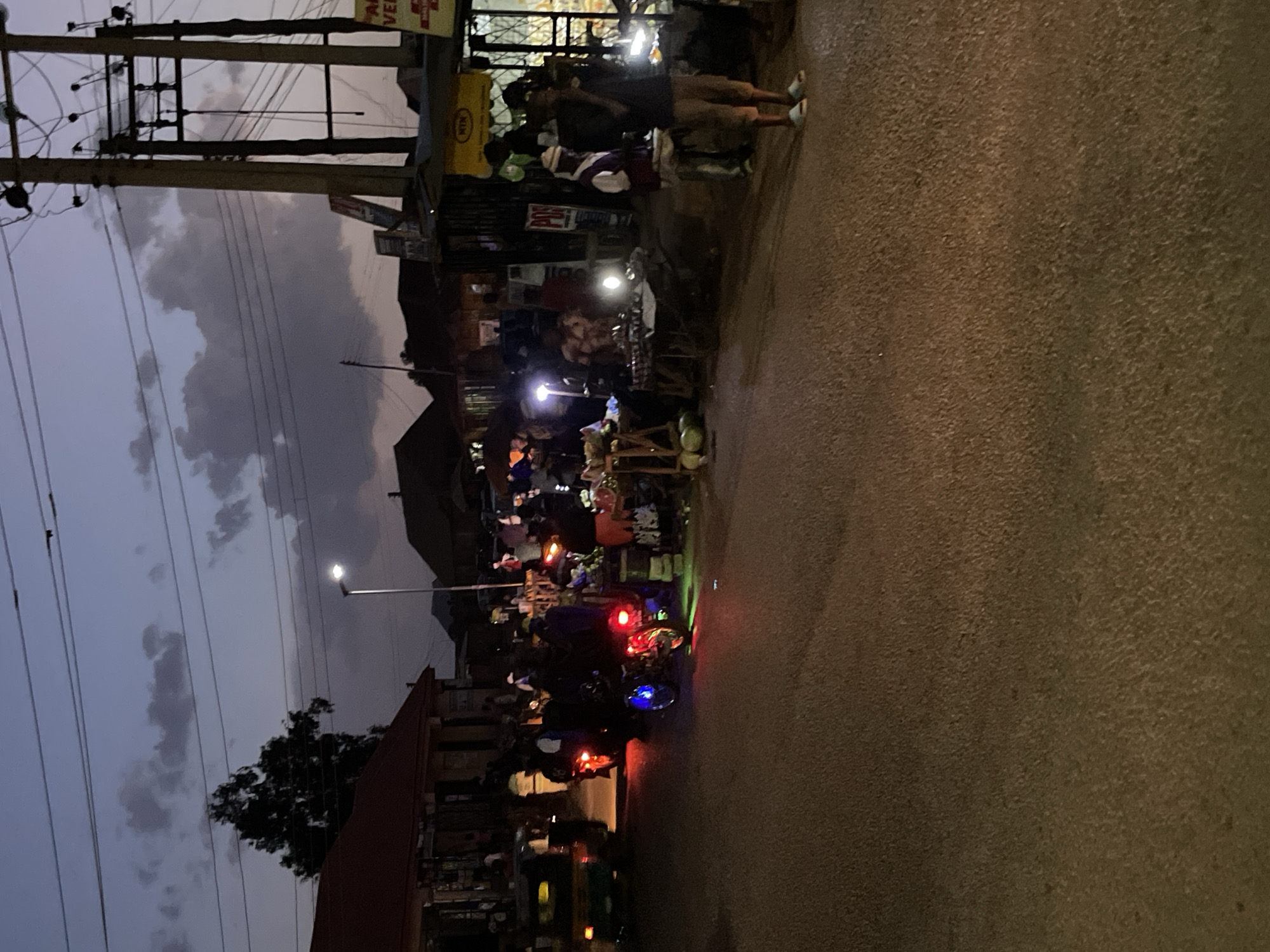 The image depicts a street scene in Bukuru, Nigeria, at dusk or night. The street is paved and dimly lit. On the left side of the image, there are utility poles with wires running overhead. A crowd of people is gathered around what appears to be a market or commercial area, likely selling food and other items. A few people are visible standing near the edge of the street. Vehicles, including motorbikes with illuminated tail lights, are present on the street. Buildings and trees are visible in the background. The sky is overcast with clouds.