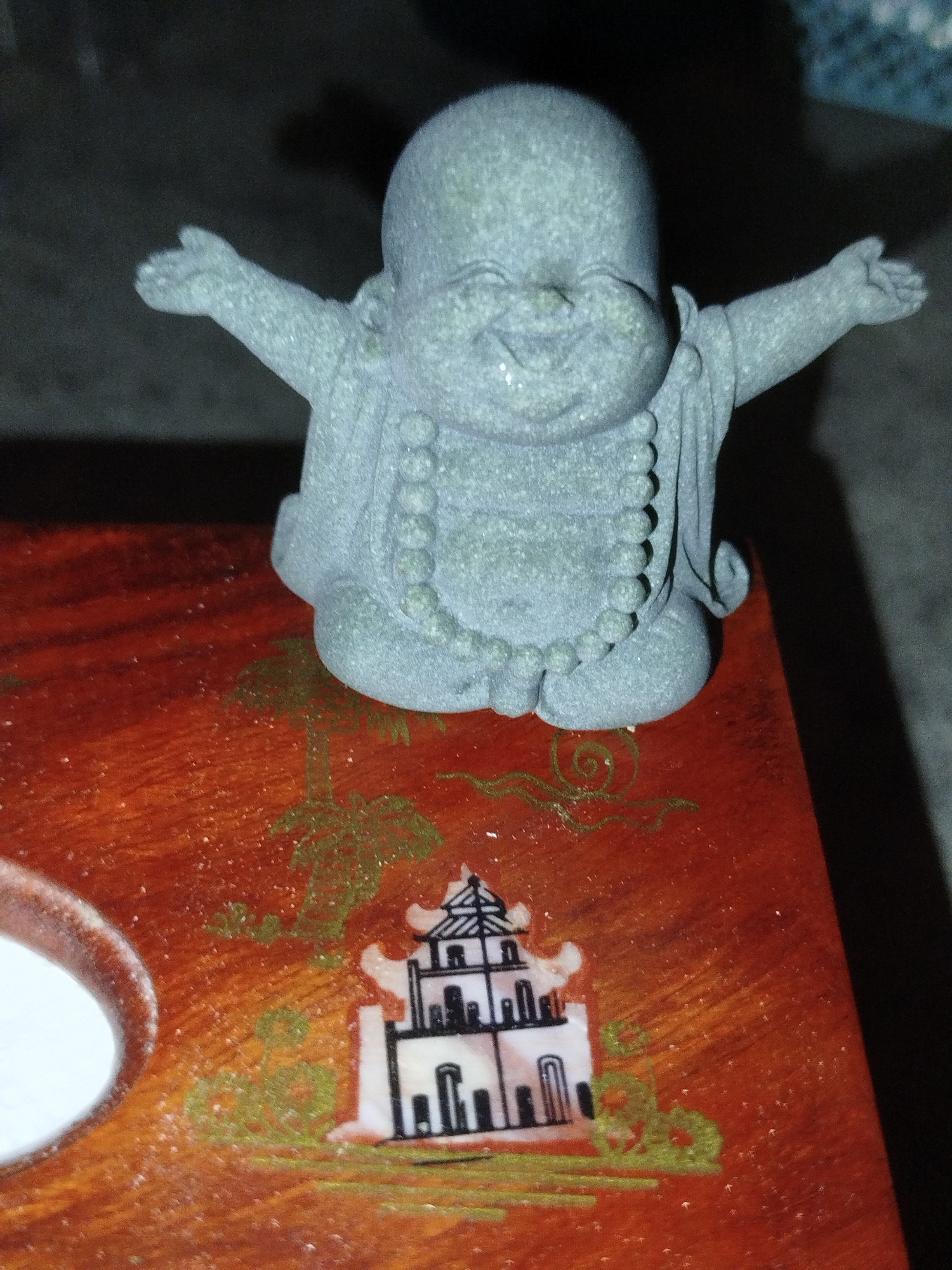 A grey, stone-like statue of a jovial Buddha with arms outstretched sits on a dark orange, lacquered wooden surface. The surface is decorated with an inlaid scene featuring a white pagoda with black architectural details, surrounded by green painted foliage. The statue is centrally placed and faces the viewer. To the left of the statue, a circular opening reveals a white interior, possibly a compartment or container. The background is dark and out of focus. The image captures an indoor setting, likely a decorative item or artifact.