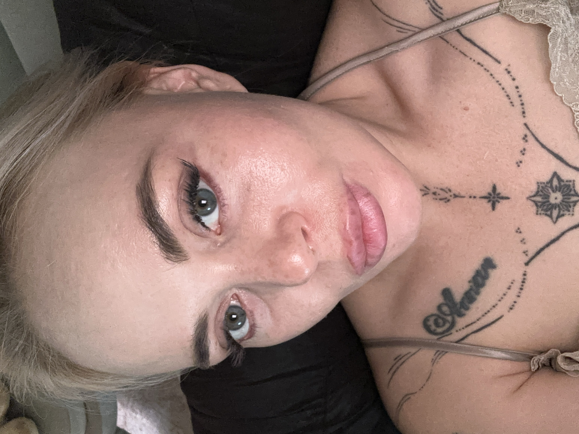 A close-up, slightly angled, eye-level, overhead shot shows a woman with fair skin and blonde hair that is pulled back, revealing her face. She is lying down and looking directly into the camera, with her head tilted slightly to her left. Her eyebrows are dark and well-defined, and she has striking blue-green eyes with visible eyelashes, including false lashes on her upper lids. Her nose is prominent and her lips are full and pink. She appears to be wearing a light-colored, thin-strapped top or dress. The skin on her face shows a visible sheen, possibly from sweat or a dewy makeup product. She has several tattoos visible on her chest and shoulder area; one on her chest reads "Amor" in cursive, and another tattoo on her sternum resembles a geometric design with a star. The background is dark and out of focus, suggesting an indoor setting. No specific location in a city is discernible from the image.